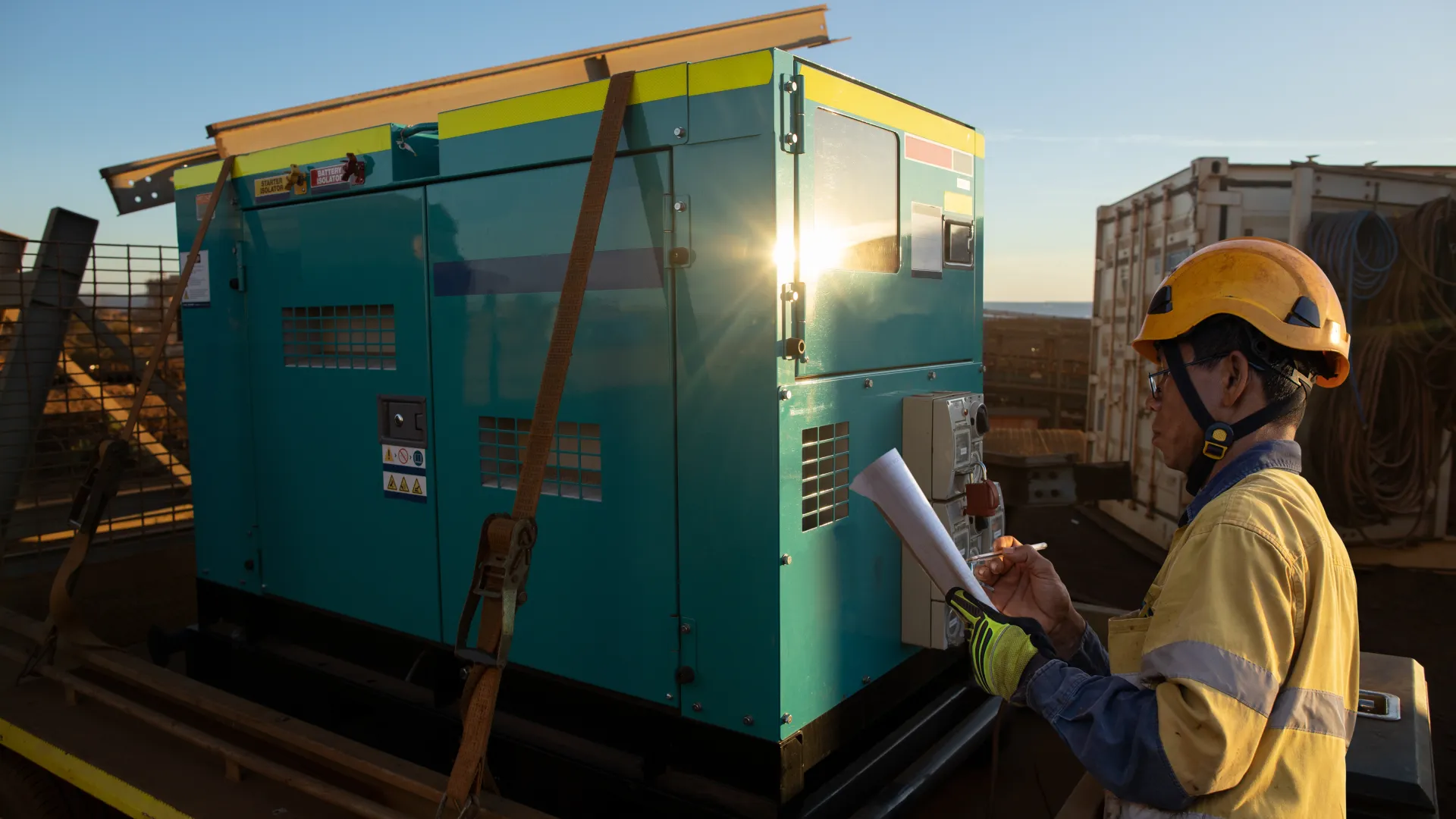 Worker reviewing details near a teal generator on a construction site at sunset.