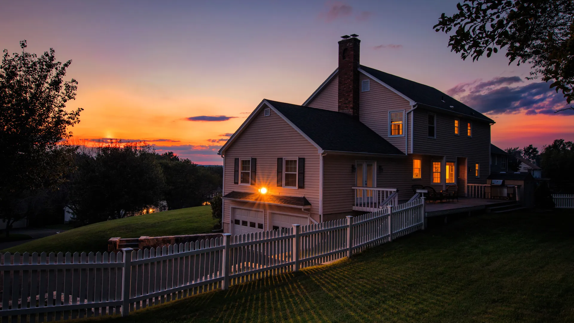 Charming two-story house at sunset, featuring a manicured lawn and white picket fence.