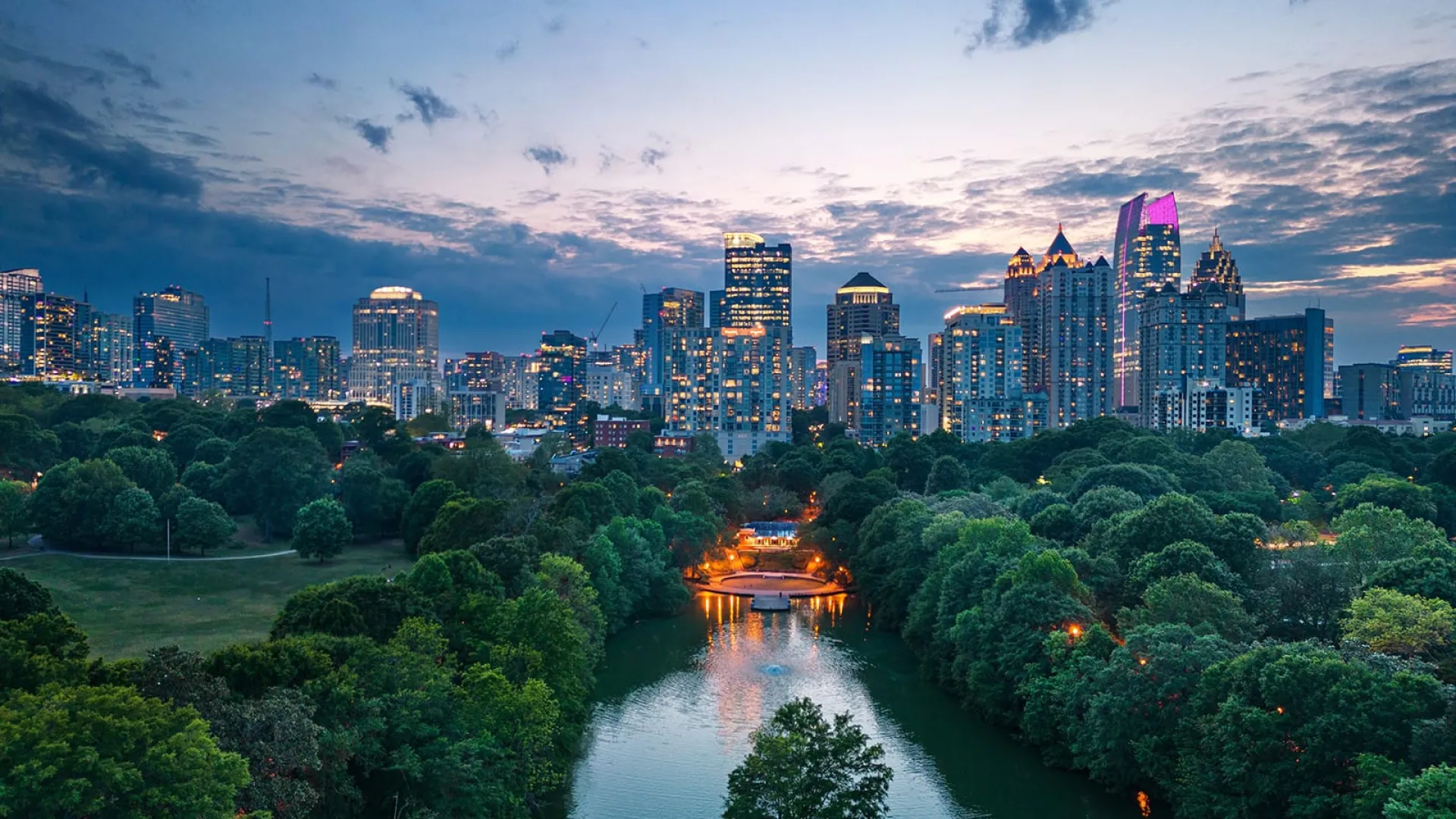 City skyline at dusk with illuminated buildings, green park, and a reflective river flowing through trees.
