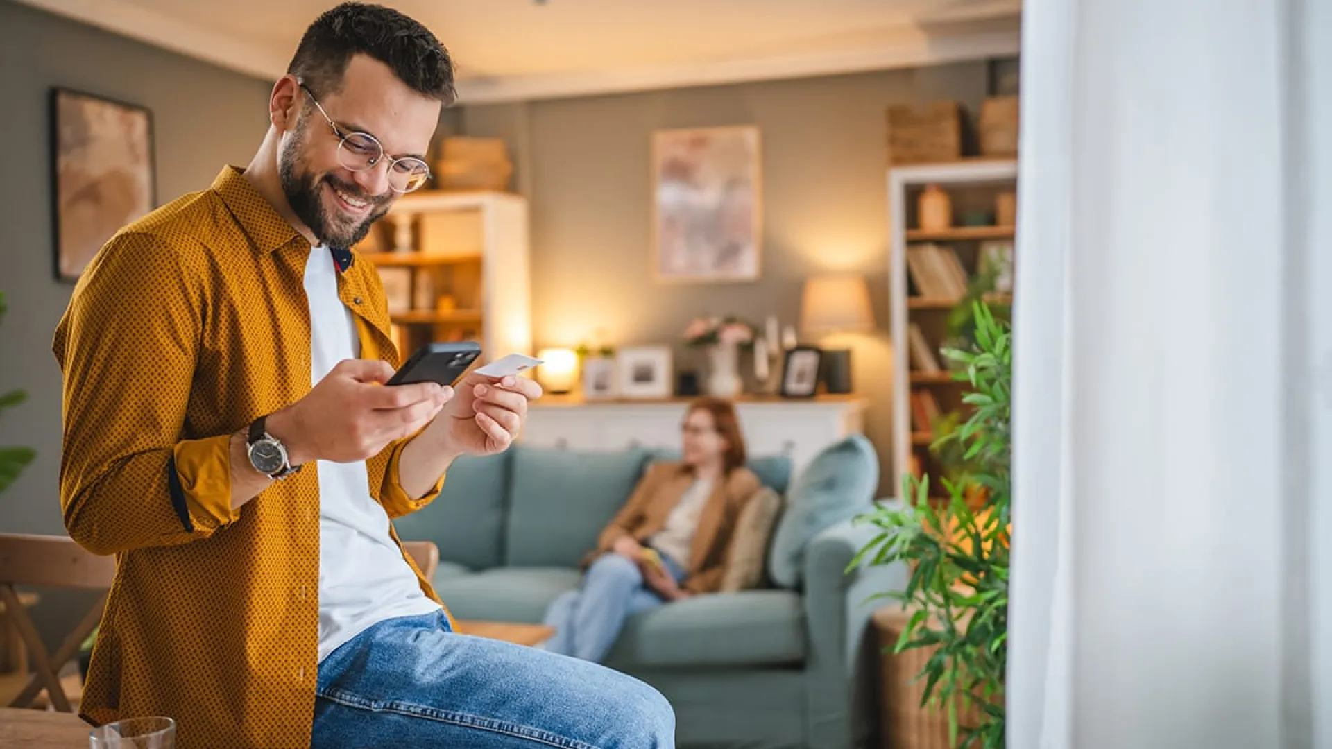 Man in casual clothes sitting on a table using smartphone while a woman relaxes on a couch in a cozy living room.