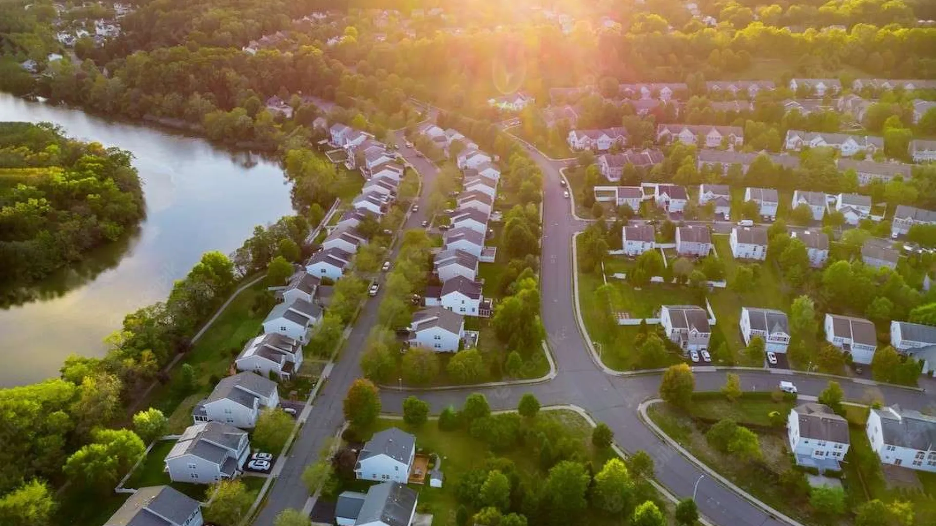 Aerial view of a suburban neighborhood with houses, trees, roads, and a river at sunset.