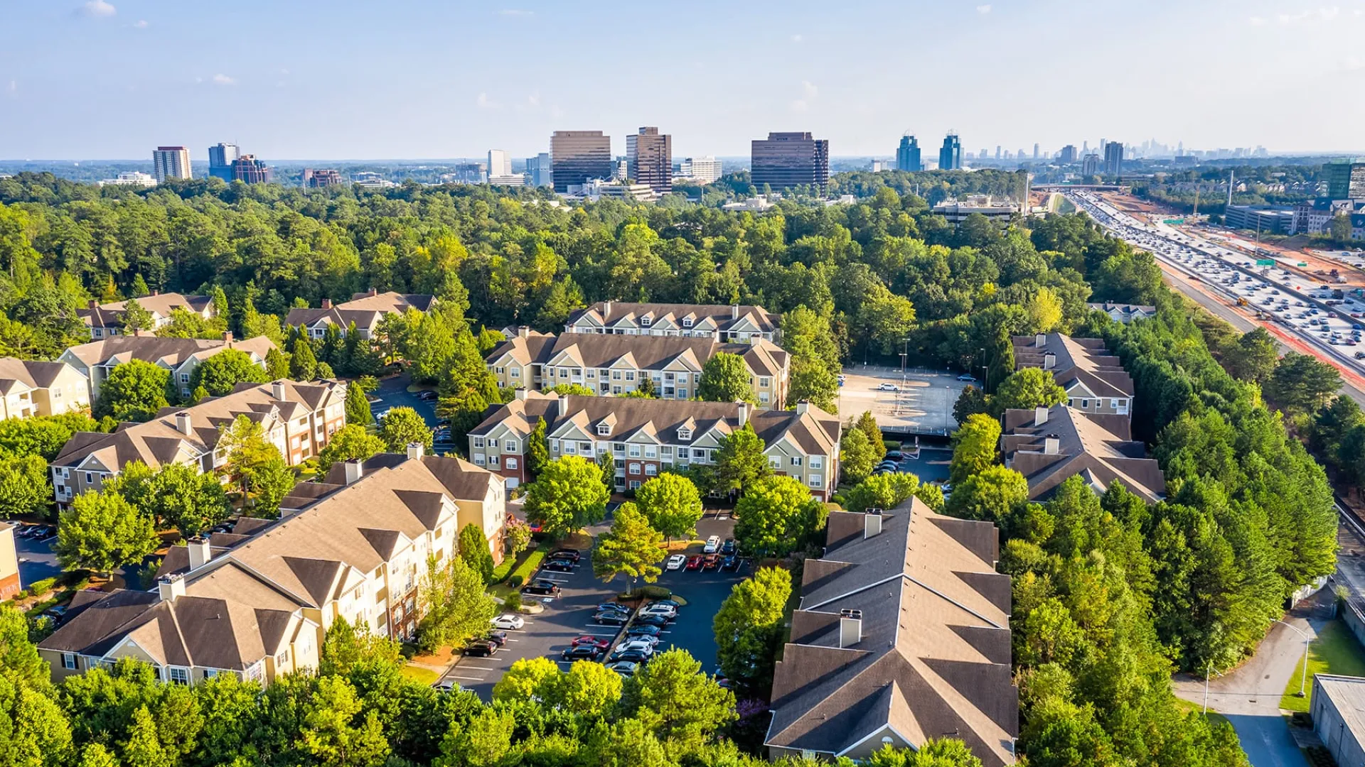 Aerial view of a suburban apartment complex surrounded by trees with a city skyline and highway in the distance under blue sky.