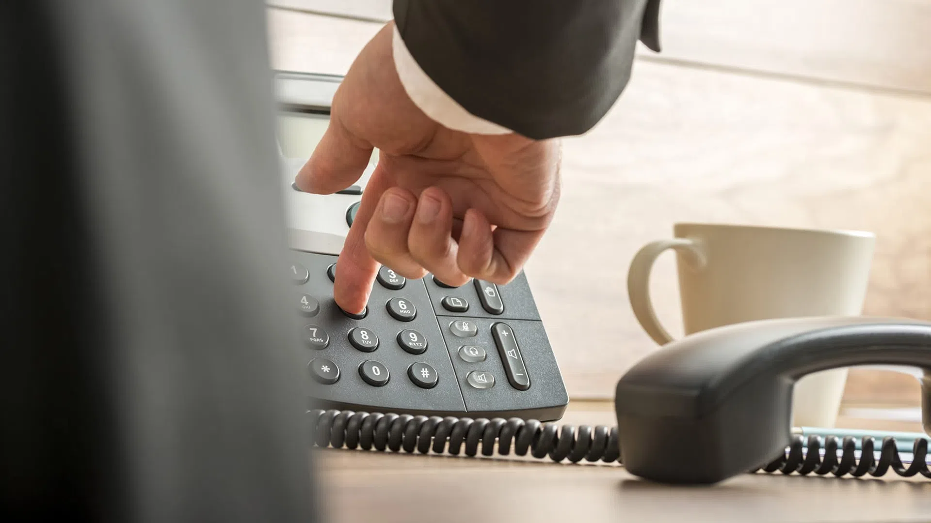 Person dialing a number on a desk phone with a coffee cup in the background in an office setting