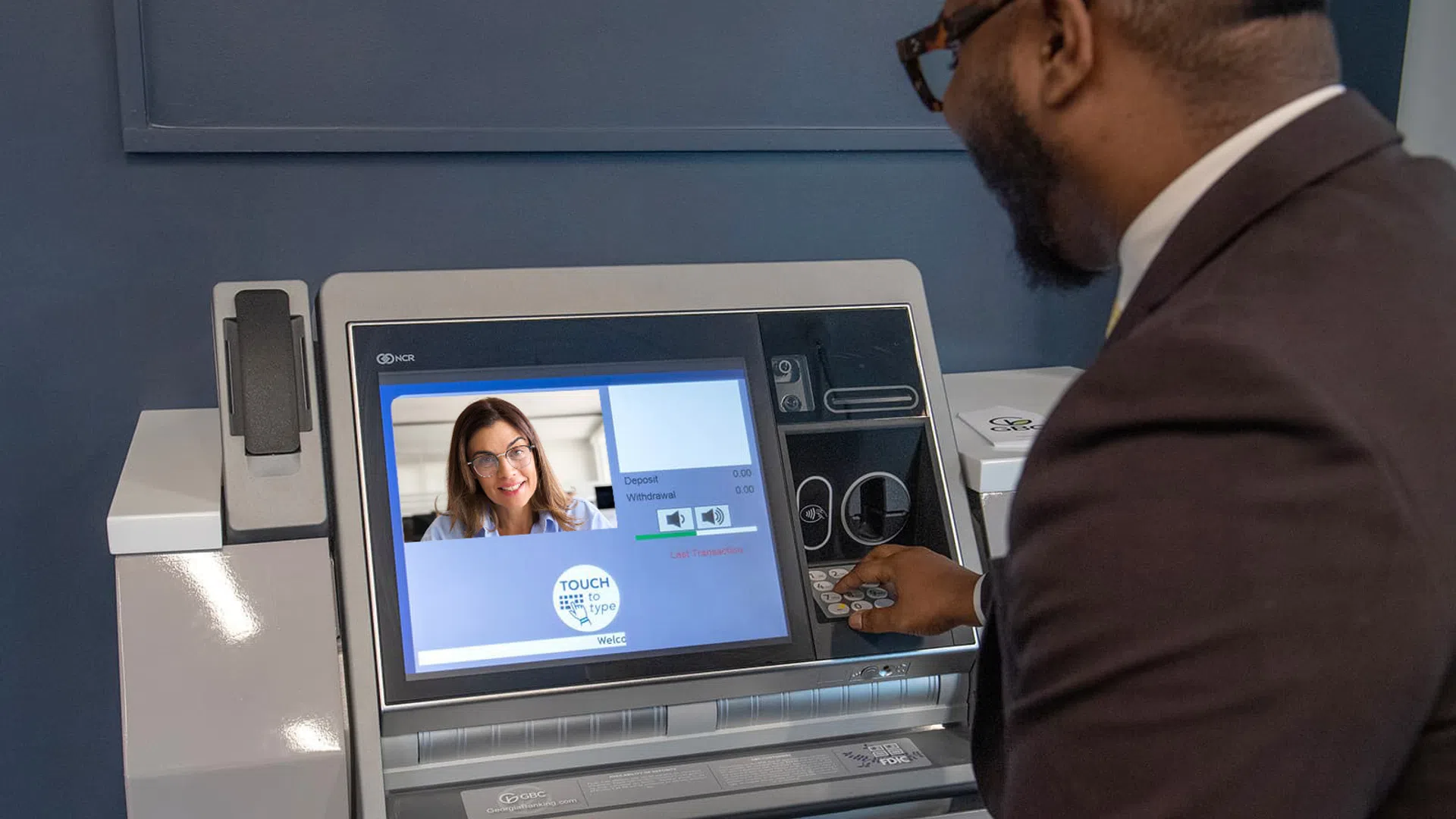 Man using video teller machine with female teller on screen for banking transaction.