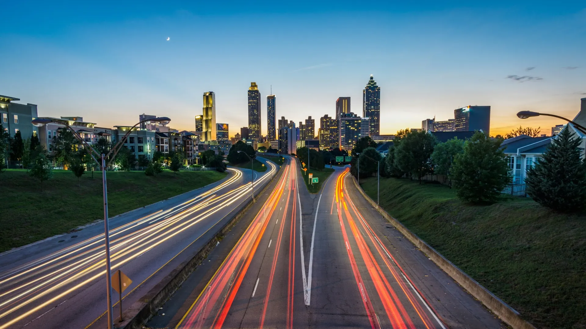 Long exposure of a highway with light trails leading to a city skyline at dusk under a clear sky.