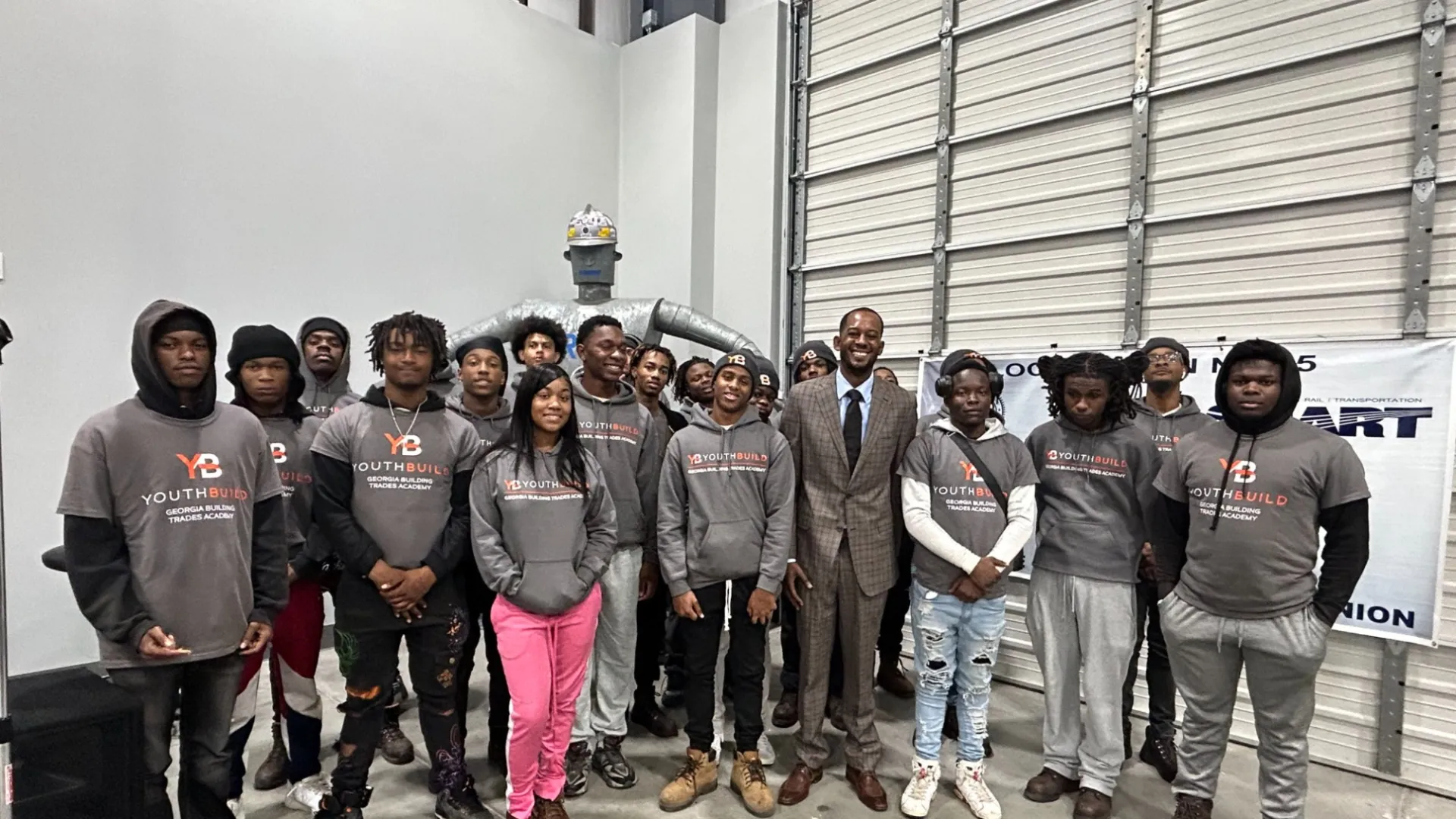 Group of young people and a man in suit posing indoors, many wearing matching YouthBuild hoodies, standing on concrete floor.
