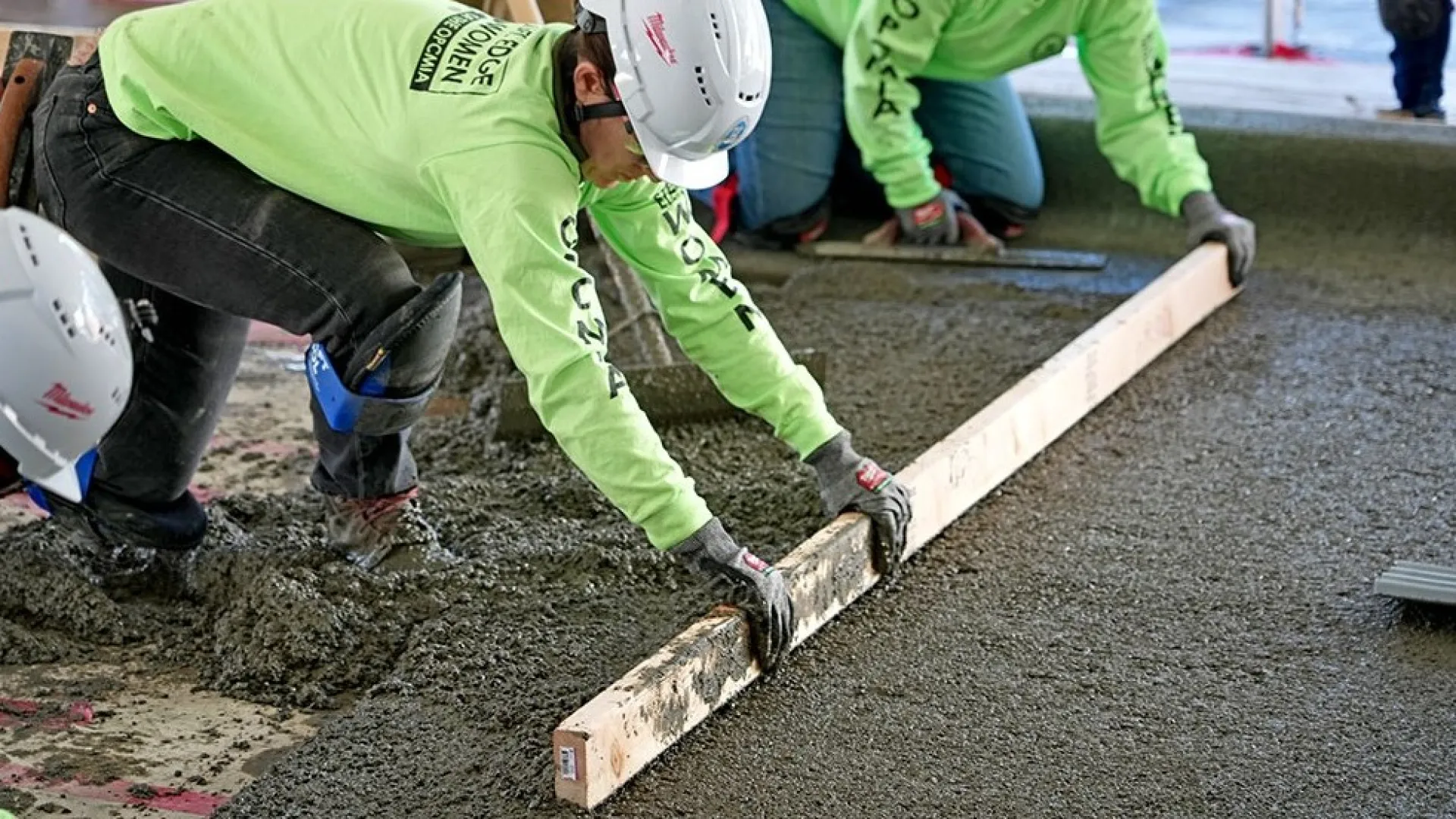 Construction workers wearing helmets and green shirts level wet concrete with a wooden screed on a building site.