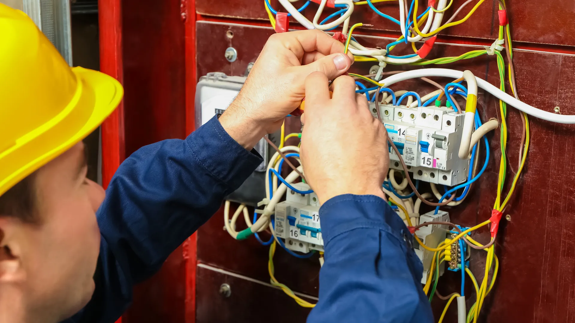 Electrician in a yellow helmet repairing colorful wiring inside an electrical panel with circuit breakers.