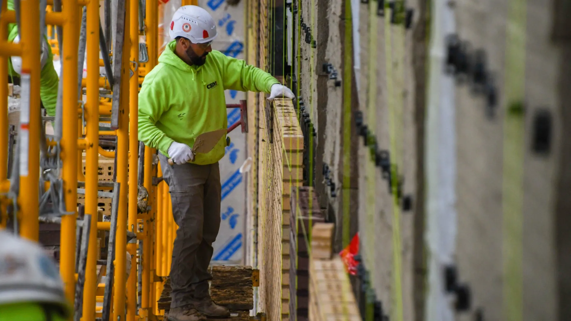 Construction worker in a green hoodie and white helmet working on brick wall with scaffolding and tools.
