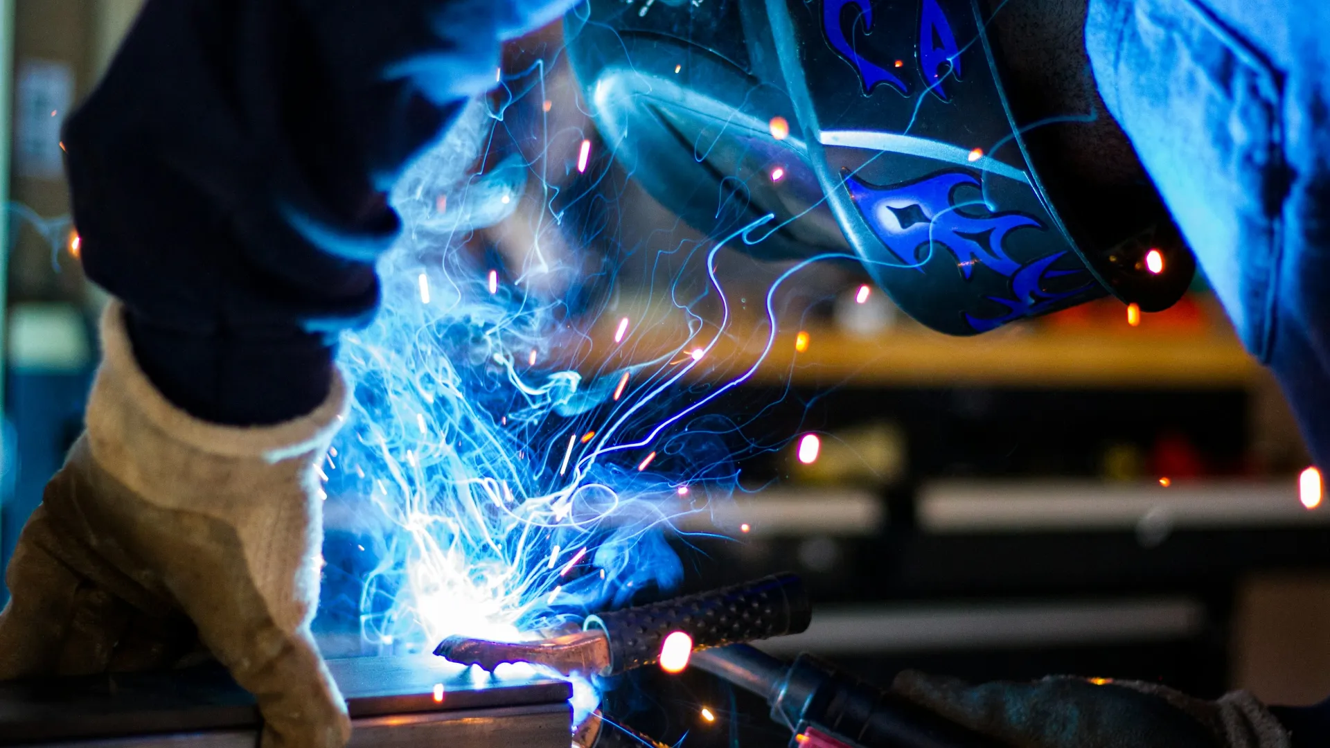 Welder in protective gear creating bright sparks while welding metal in a workshop setting.
