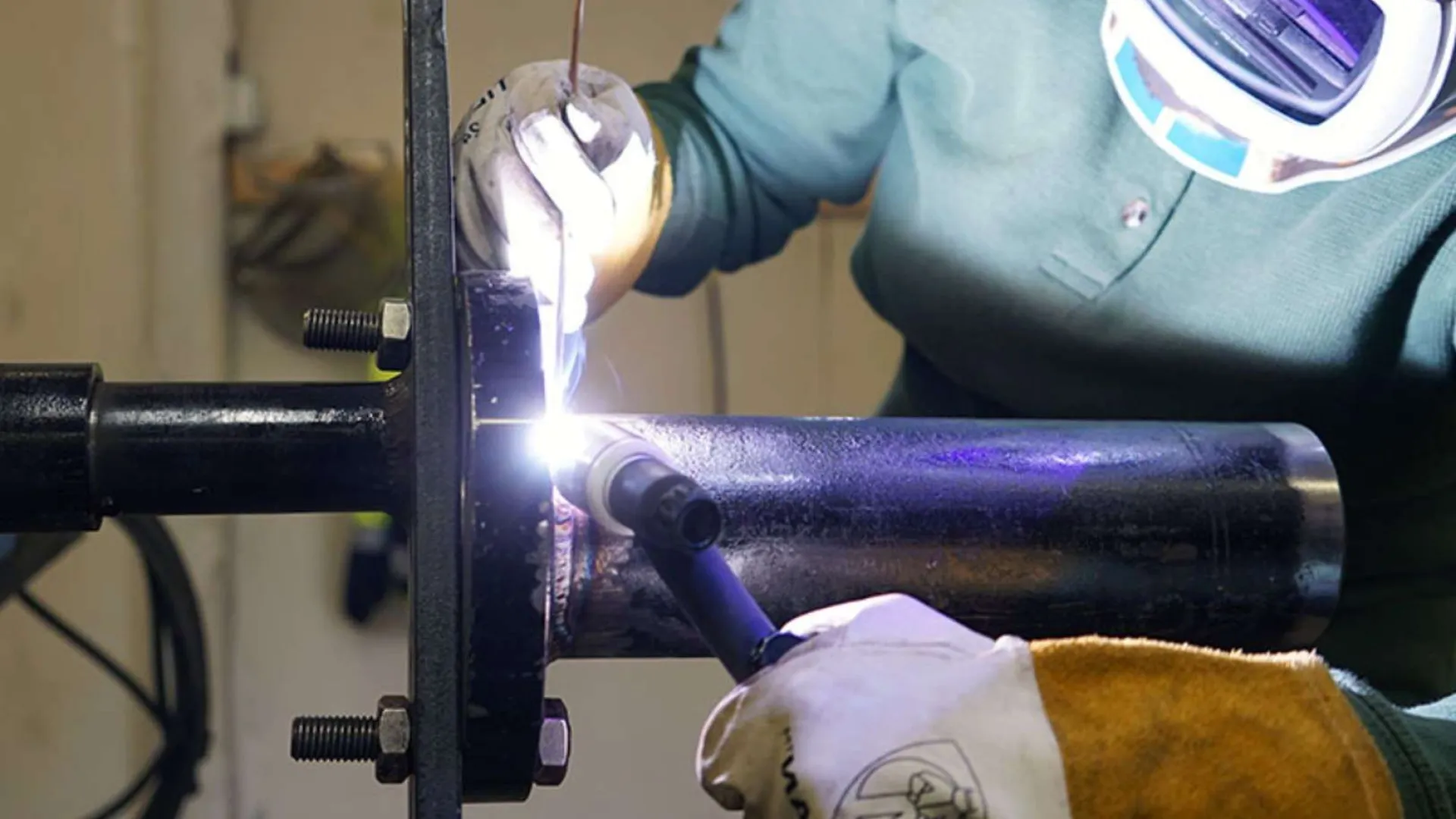 Welder wearing protective gloves and helmet welding a metal pipe with sparks flying.