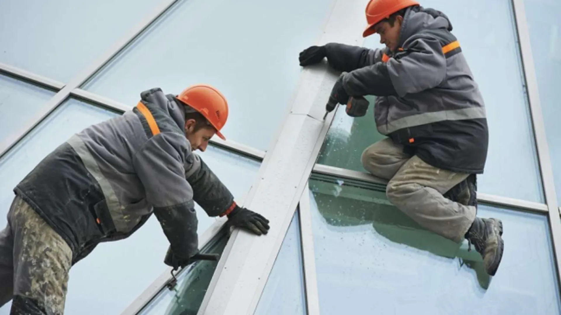 Two construction workers in safety gear installing a large window panel on a building exterior.