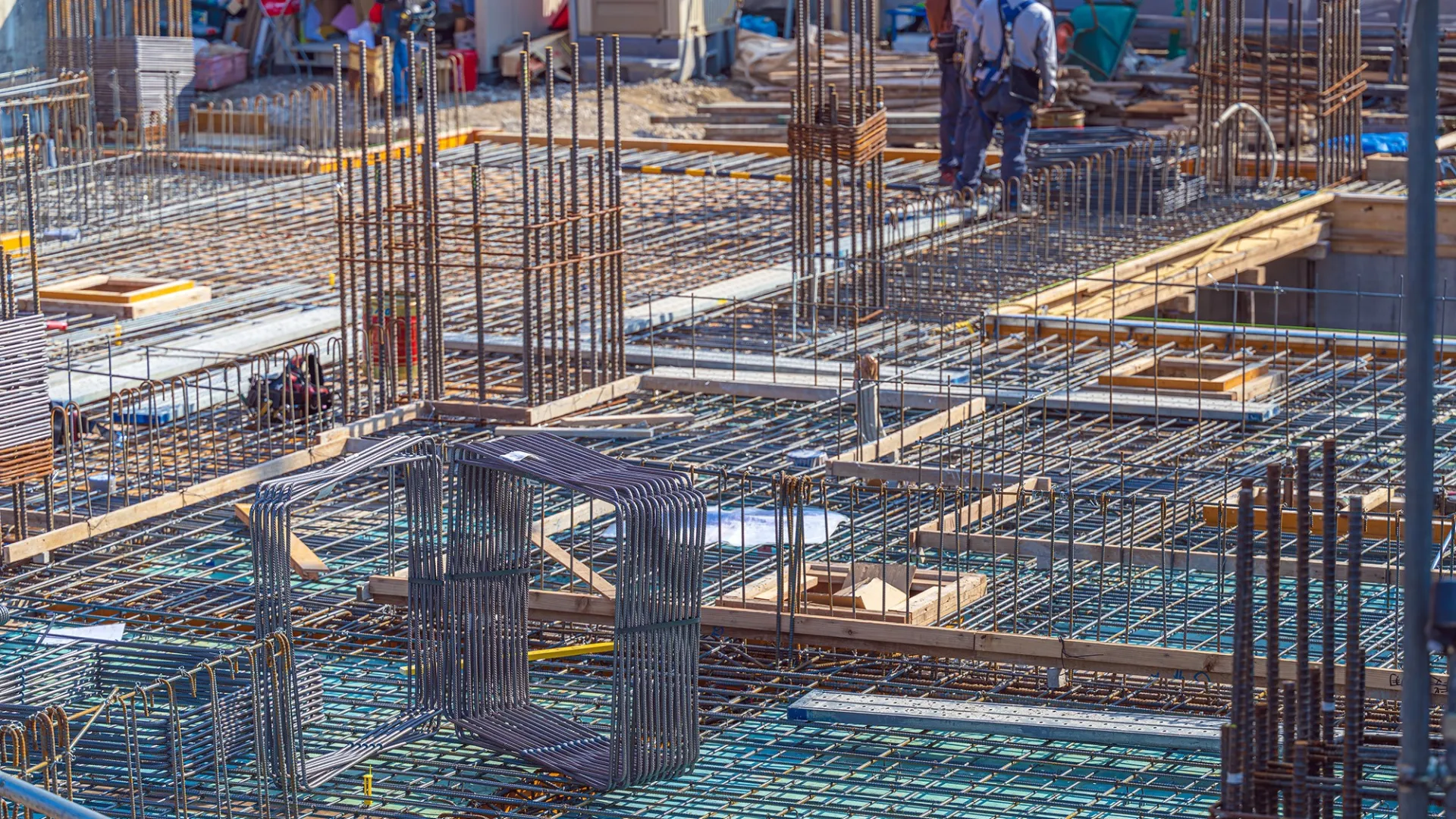 Steel rebar framework for concrete foundation at a construction site with workers in the background.