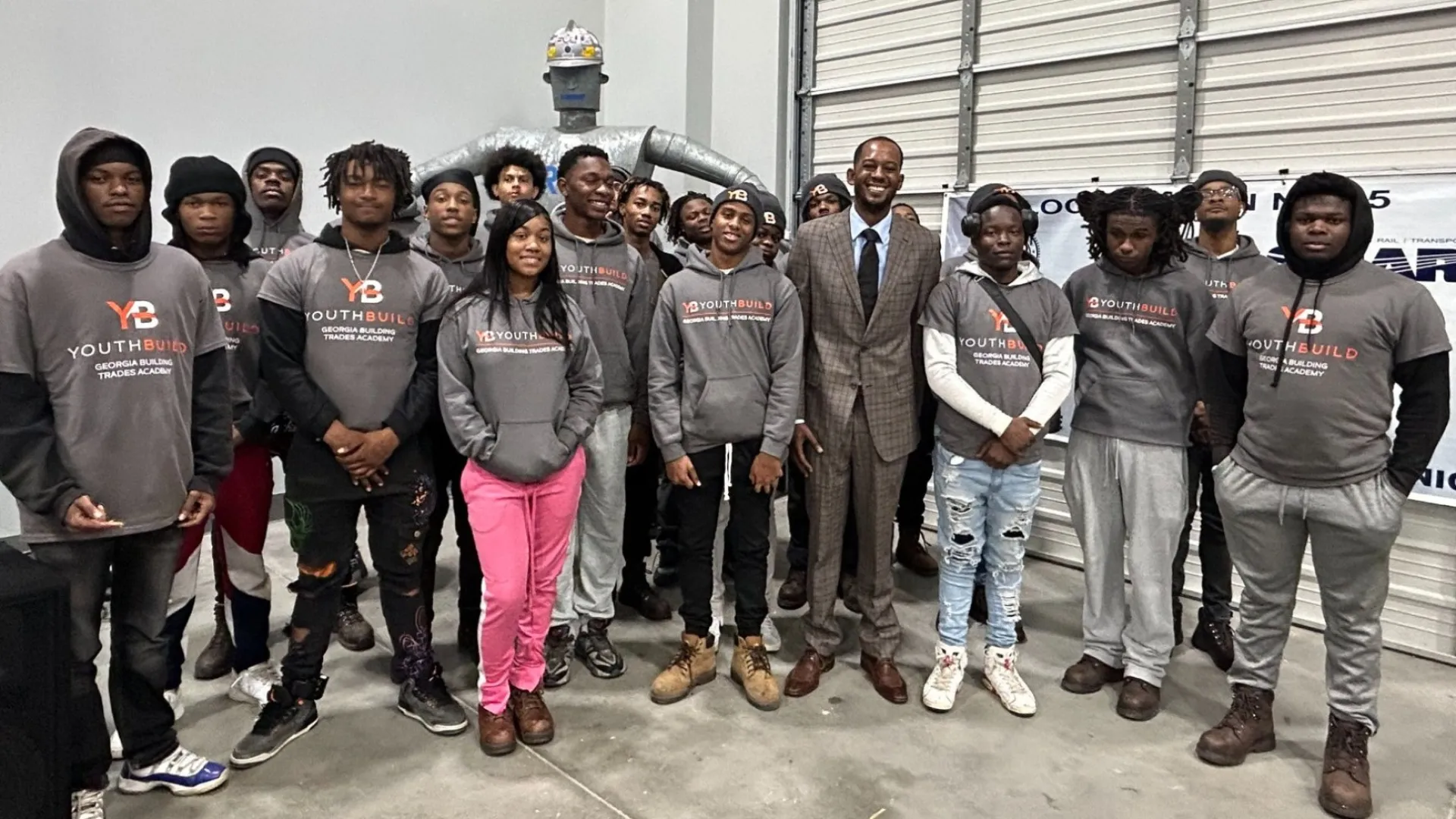 Group of young people and a man in suit posing indoors, many wearing matching YouthBuild hoodies, standing on concrete floor.