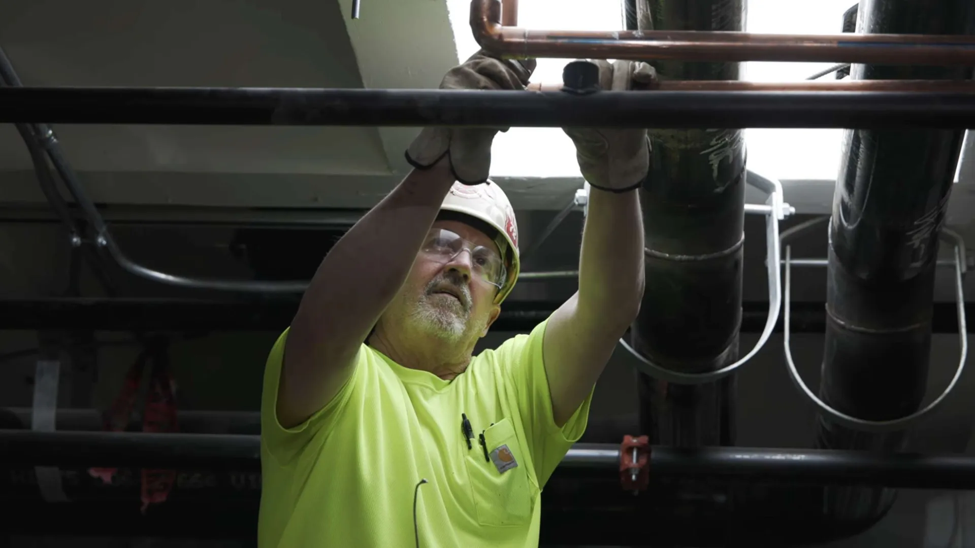 Construction worker wearing safety glasses and helmet installs or repairs pipes overhead in industrial setting.