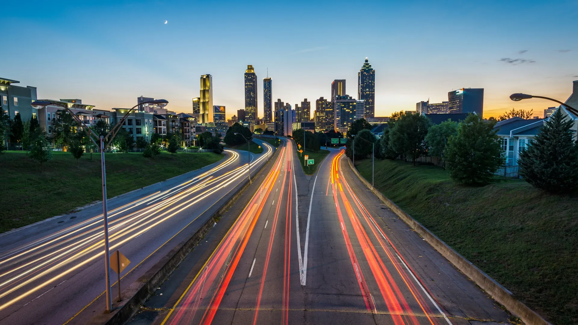 City skyline at dusk with highway light trails leading towards tall buildings under a clear sky.