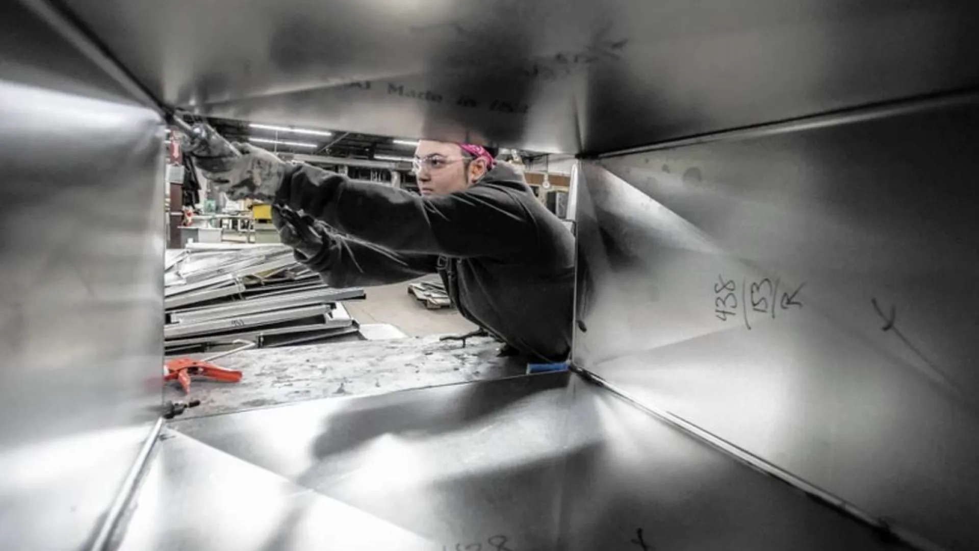 Worker assembling metal ductwork in a factory seen through a rectangular metal frame with industrial tools nearby.