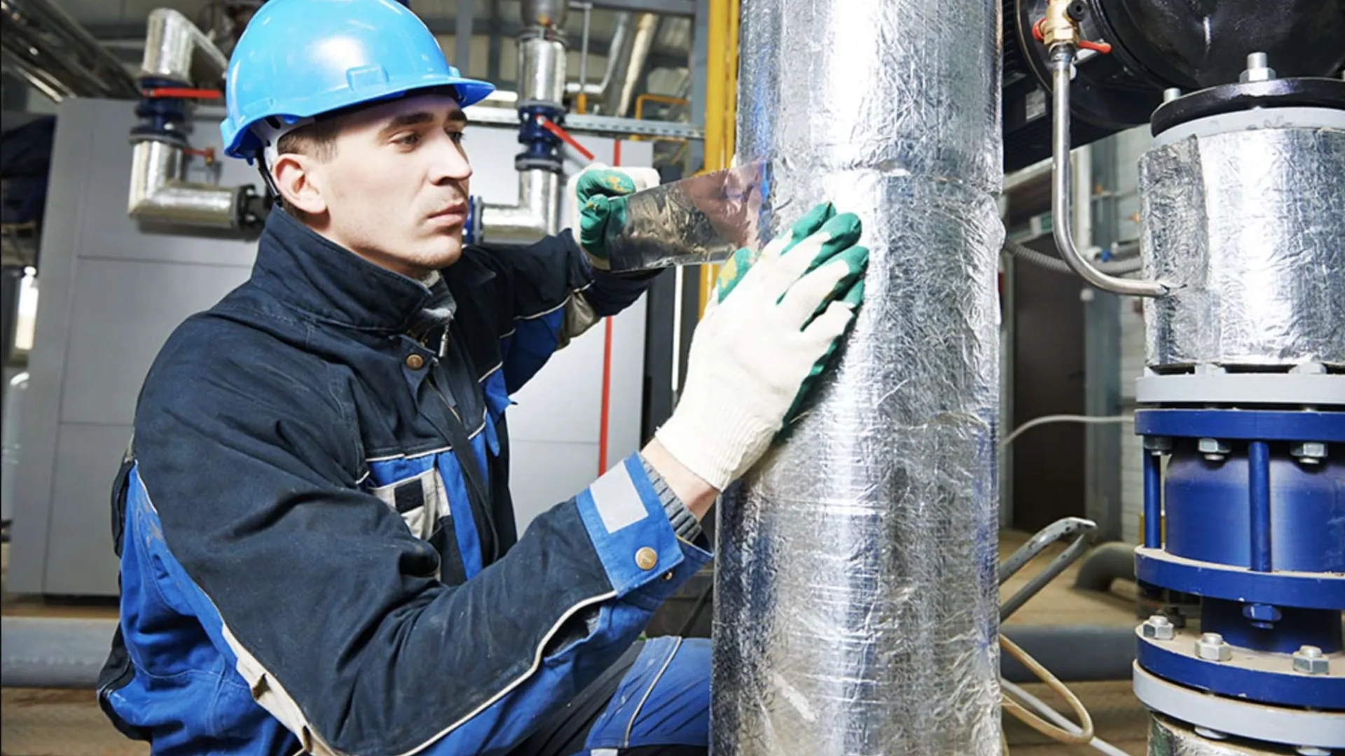 Worker in blue helmet and uniform applying insulation tape on industrial pipe in factory setting.