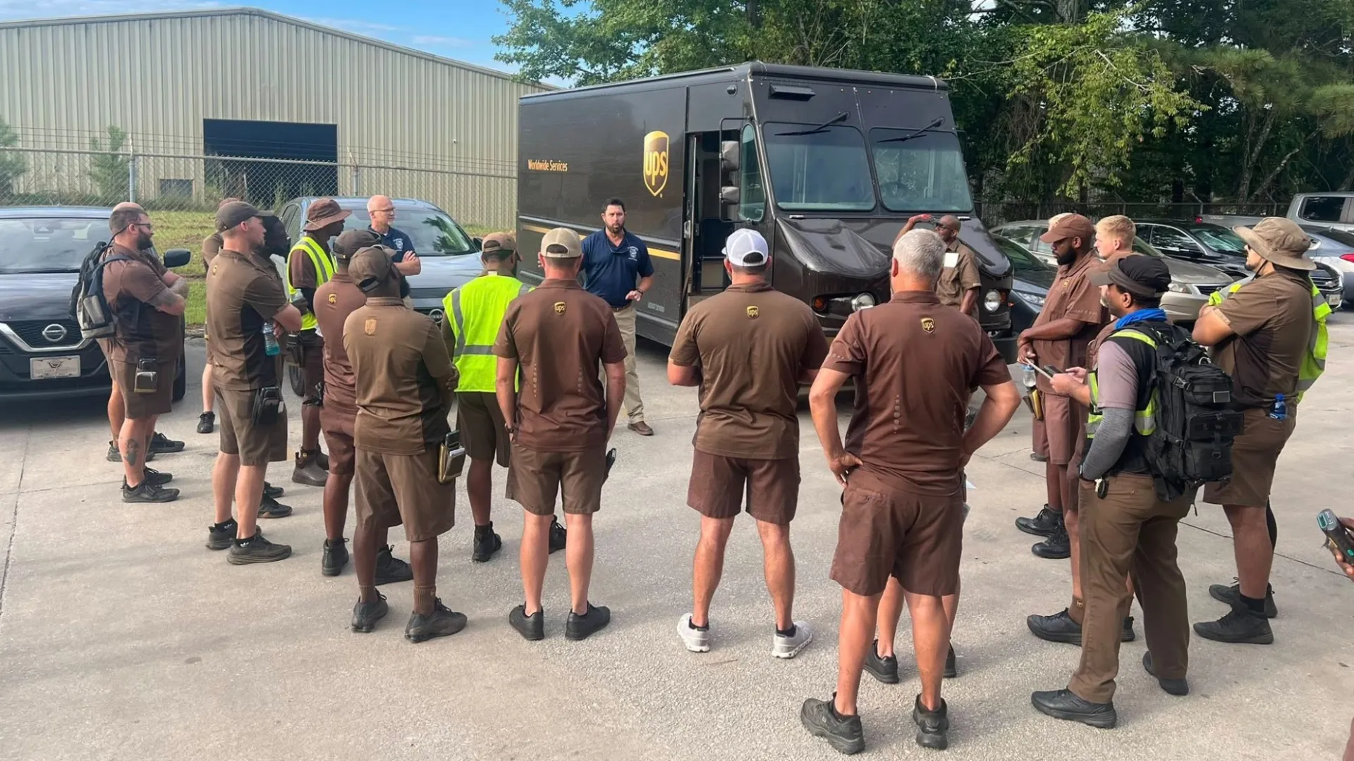 Group of UPS delivery drivers in uniform gathered outdoors next to a UPS truck during a briefing.