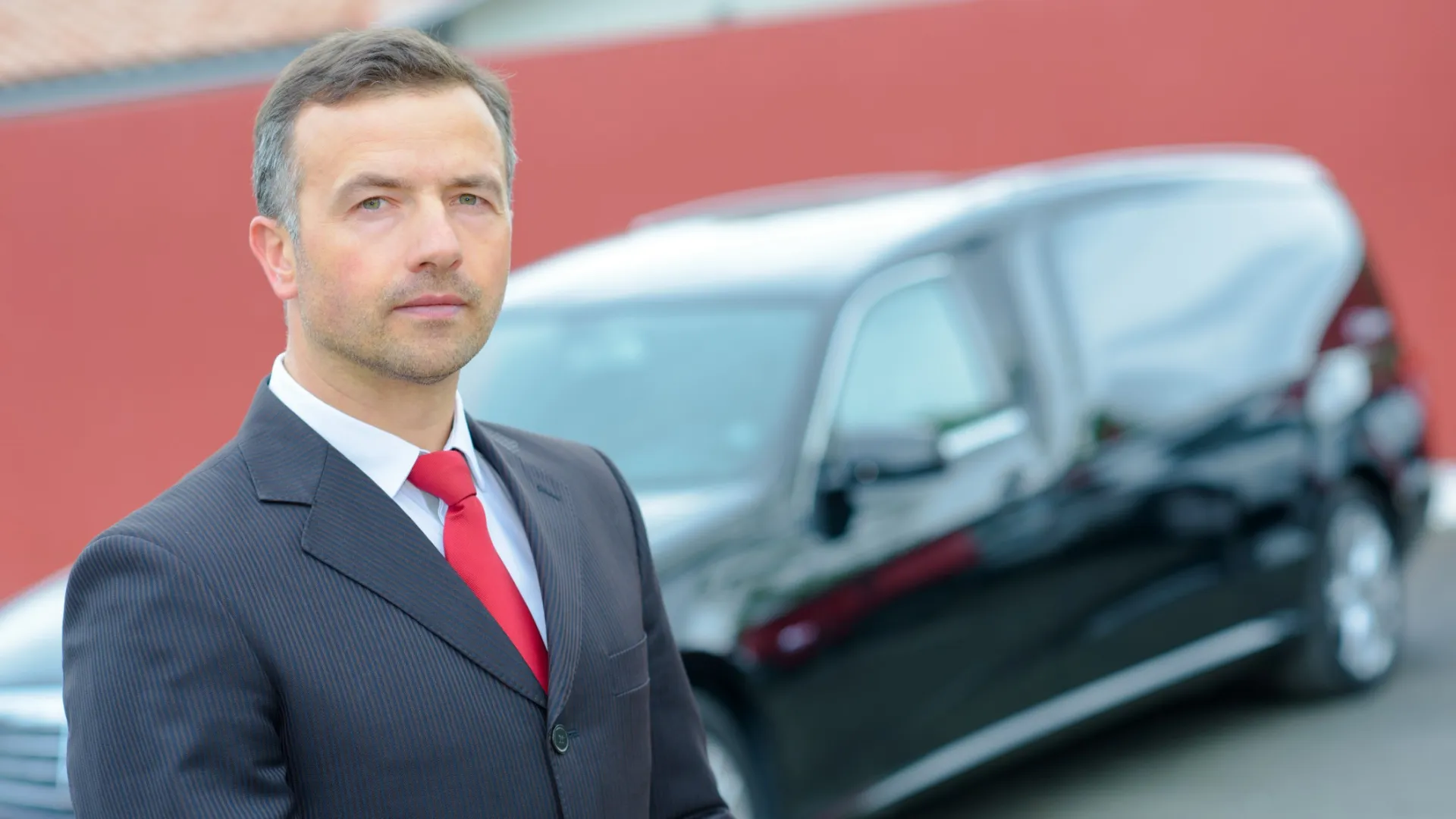 Confident man in suit and red tie standing next to a black luxury car with a red wall background.