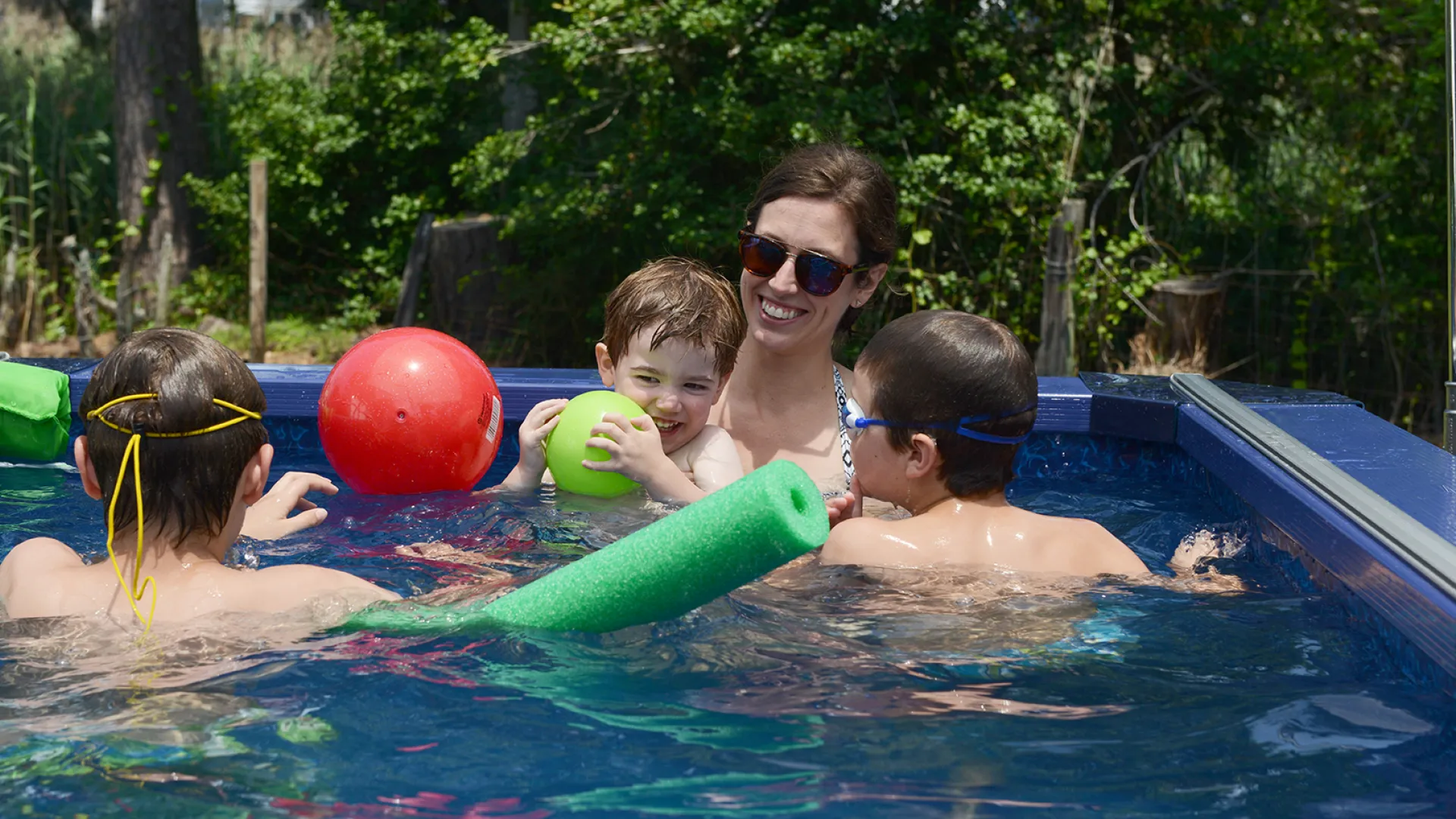 Mother and three children playing with balls and pool noodles in an outdoor swimming pool on a sunny day