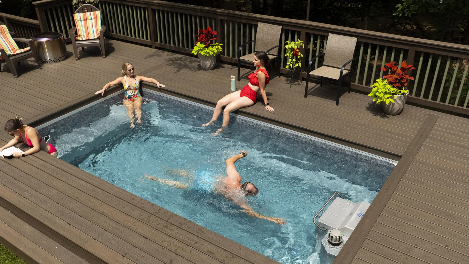 Four people enjoying a built-in backyard swimming pool with wooden deck and outdoor seating.