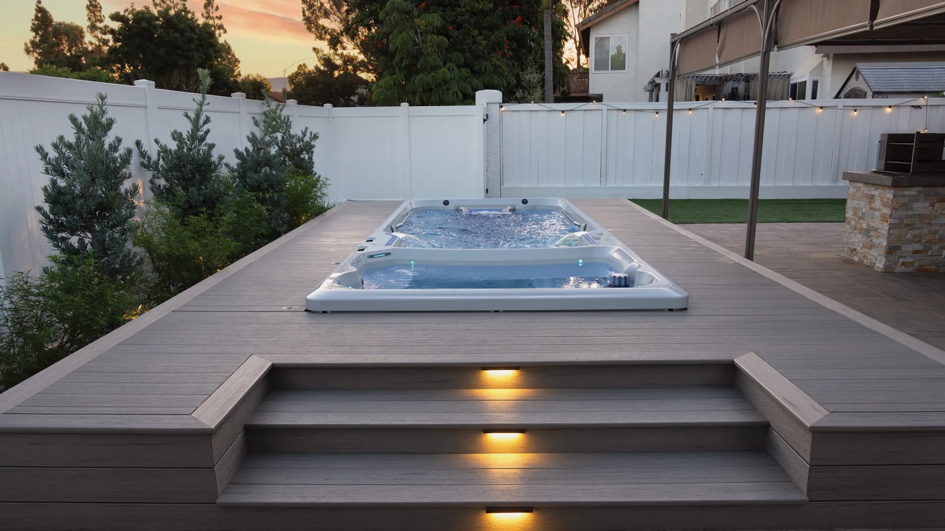 Modern backyard swim spa built into a wooden deck with stairs and ambient lighting at sunset.