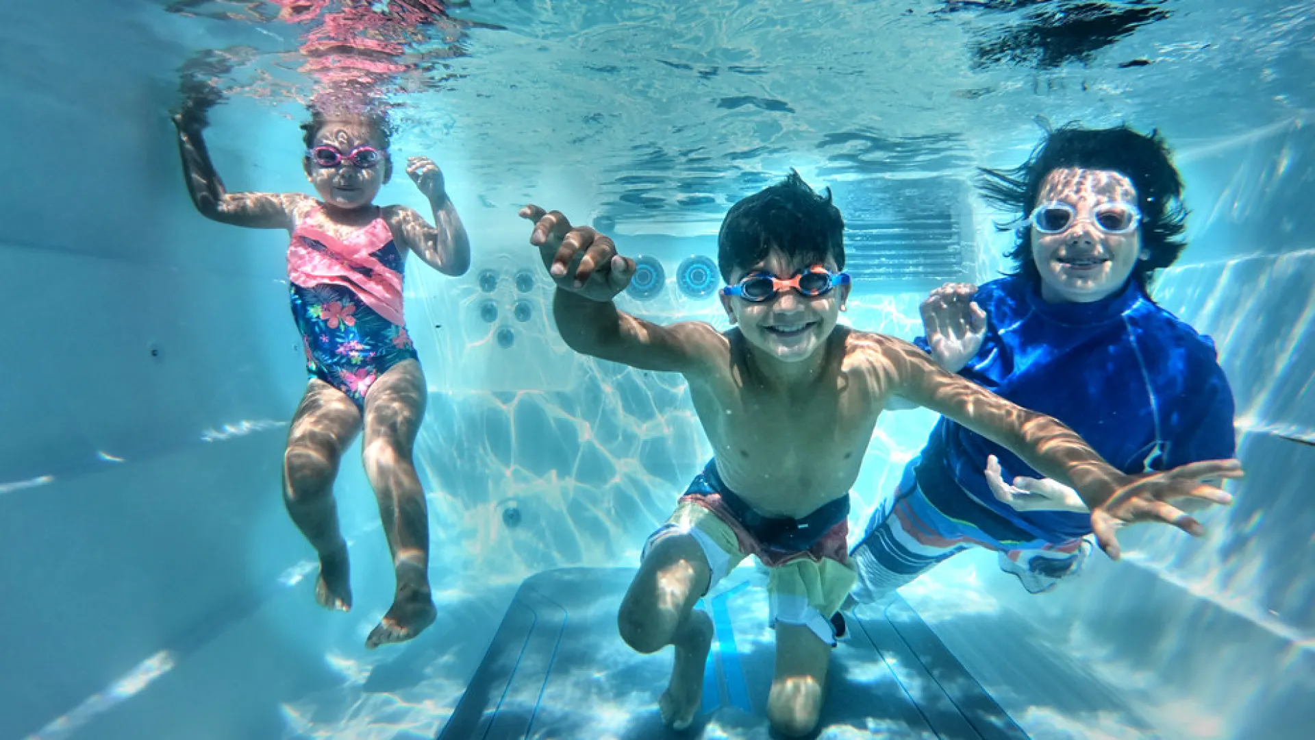Three children wearing goggles swimming underwater in a clear pool, smiling and reaching toward the camera.