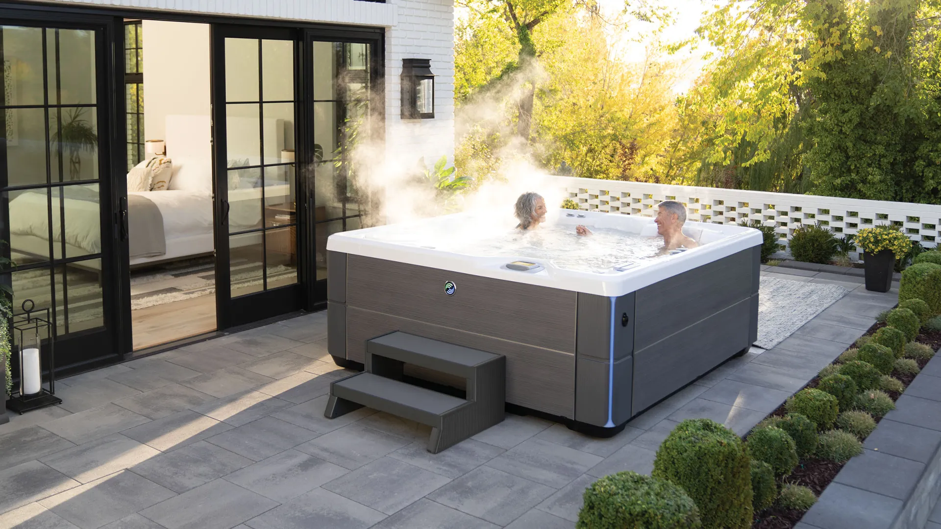 Couple enjoying a steaming outdoor hot tub on a patio next to a modern house with glass doors.
