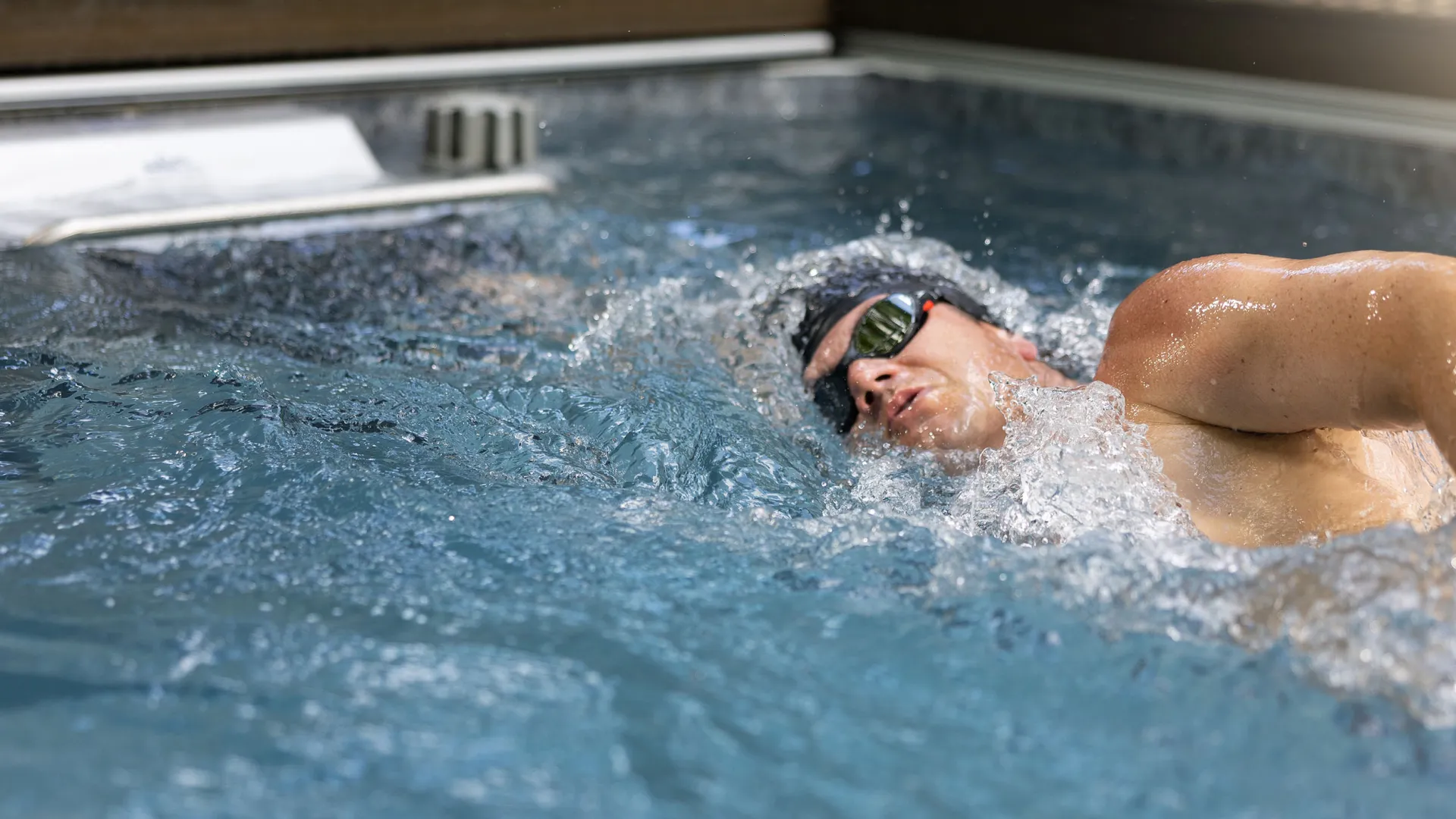 Male swimmer wearing goggles swimming freestyle in an indoor pool with splashing water and bright lighting.
