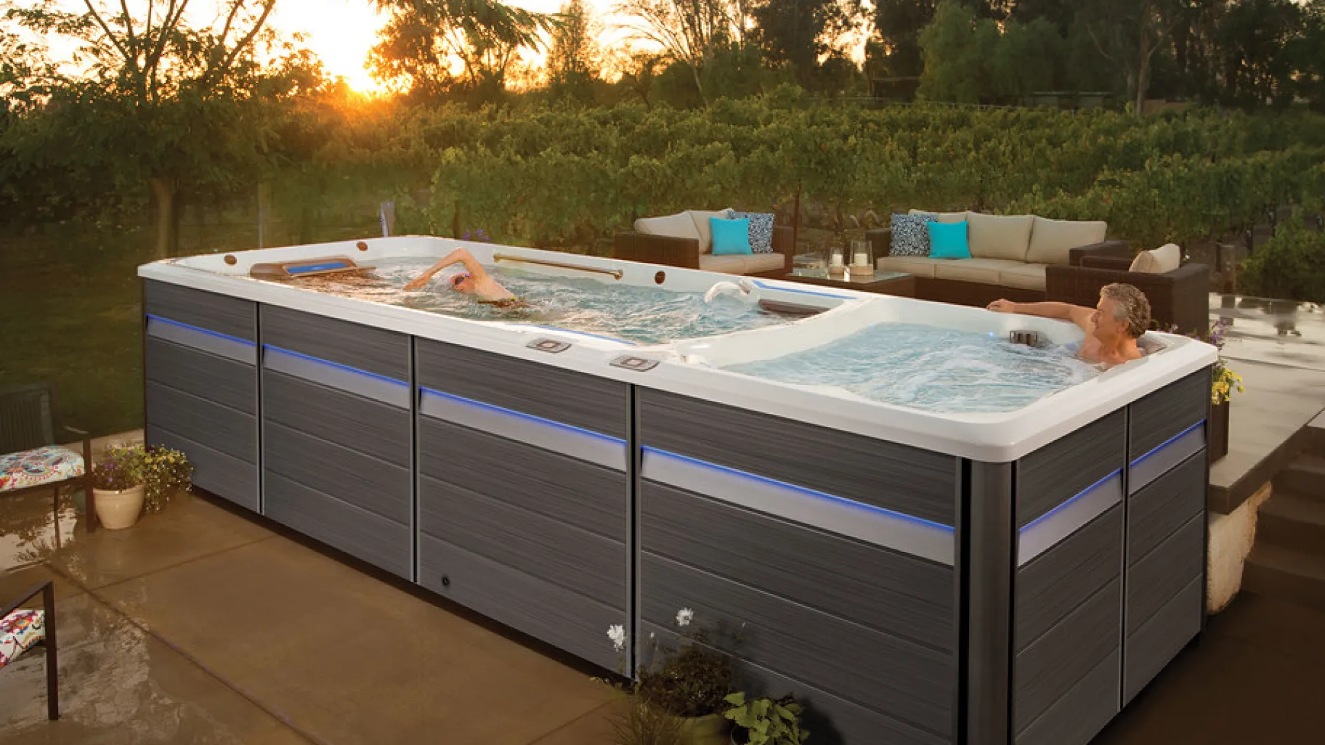 Modern outdoor swim spa with two people swimming and relaxing in a garden setting at sunset.