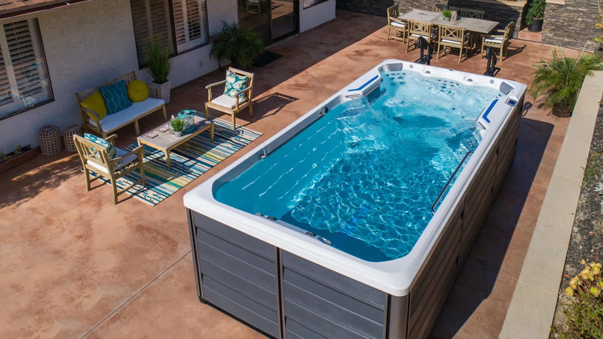 Backyard swim spa with clear blue water and adjacent patio furniture on concrete deck under natural light