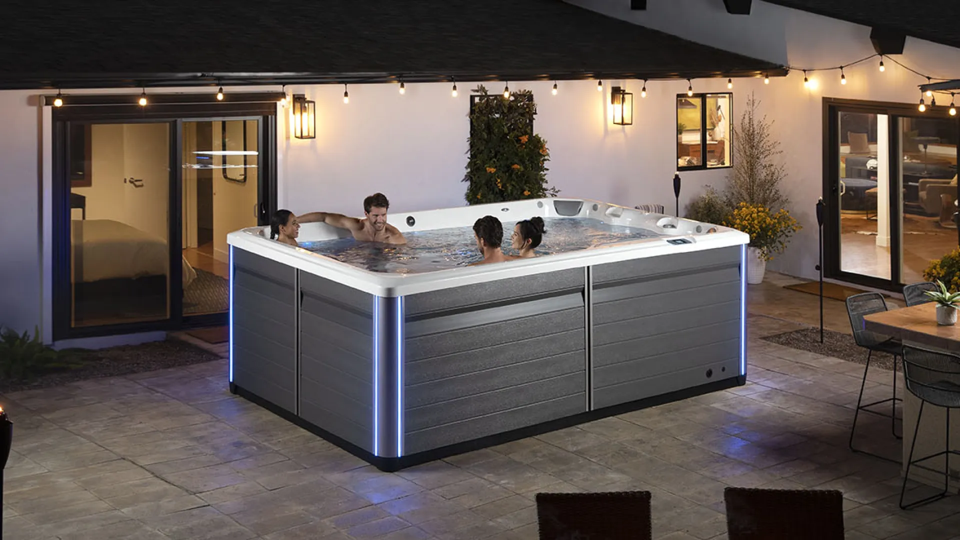 Four people enjoying a modern outdoor hot tub on a patio at night with ambient string lights and seating area.