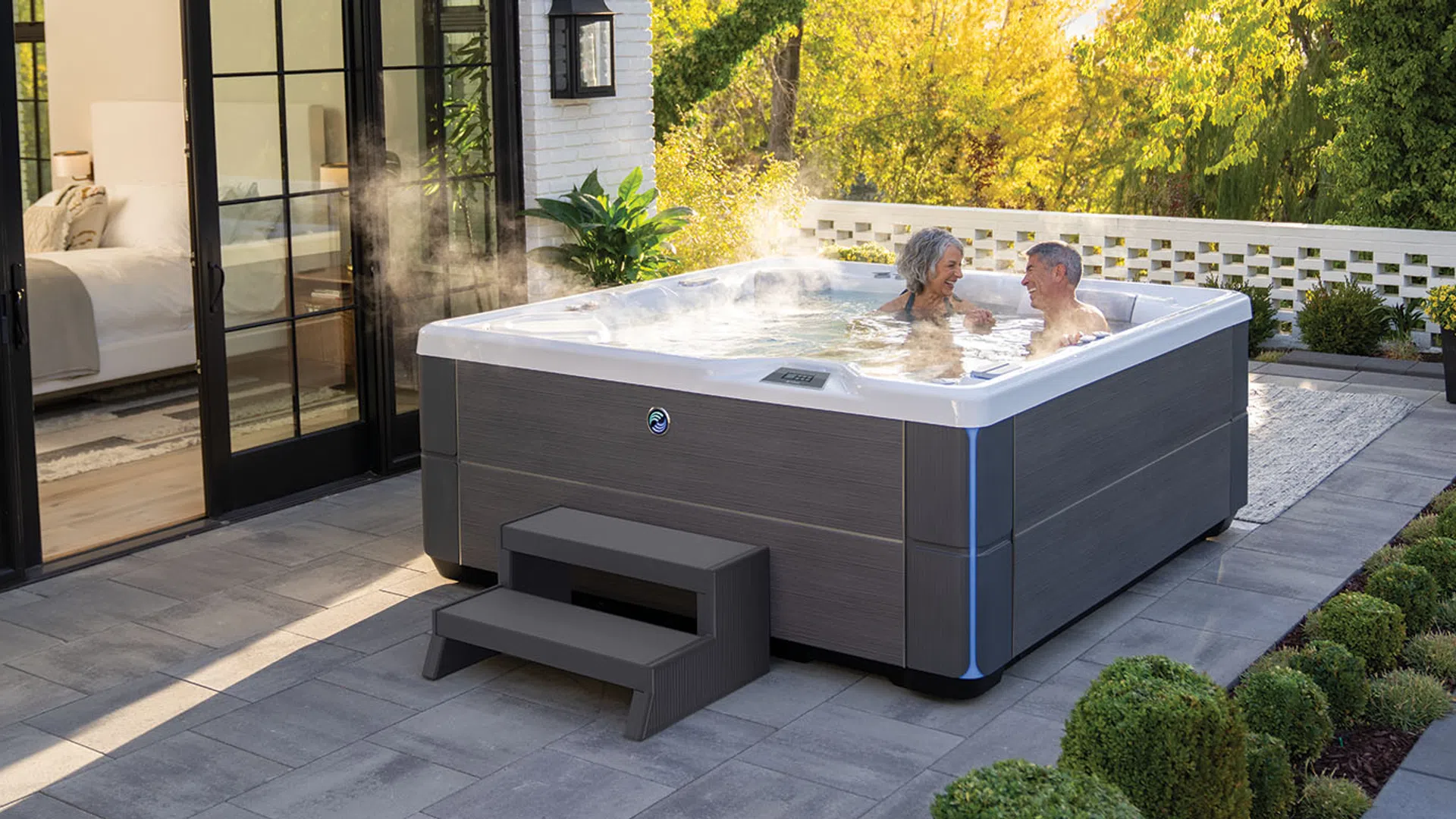 Couple relaxing in a modern outdoor hot tub on a patio surrounded by greenery and modern house windows.