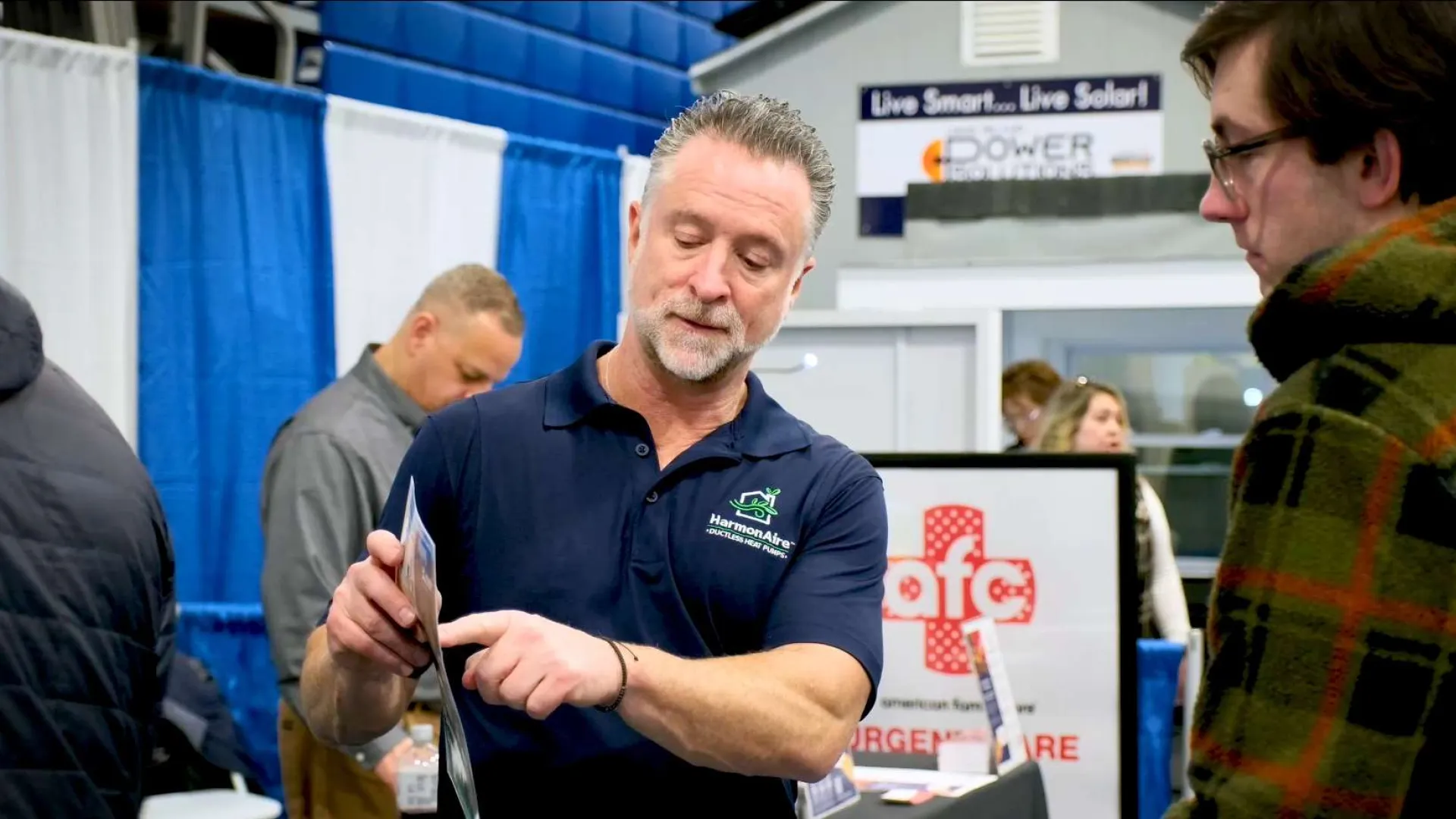 Man in navy blue polo explaining product to visitor at a trade show booth with informational displays.