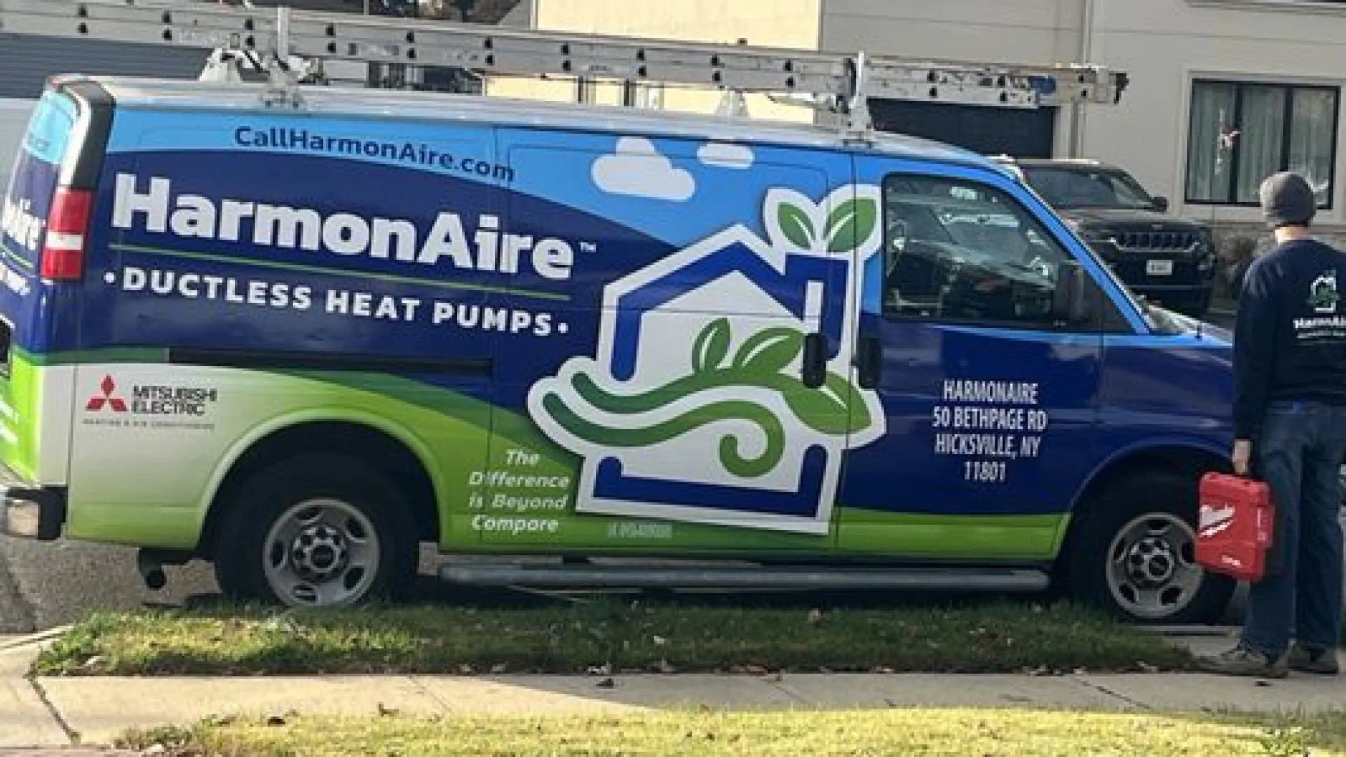 HarmonAire ductless heat pumps service van parked on street with technician holding red toolbox.