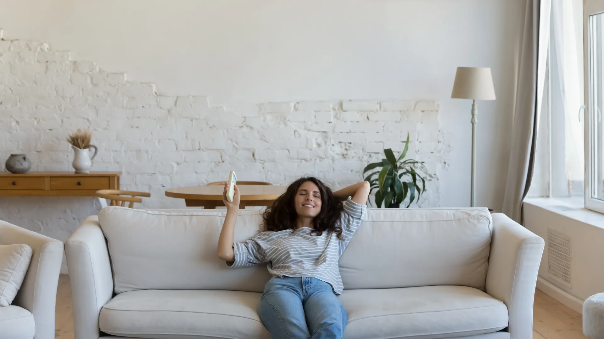 Woman relaxing on a white couch under a wall-mounted air conditioner in a bright living room with plants and decor.