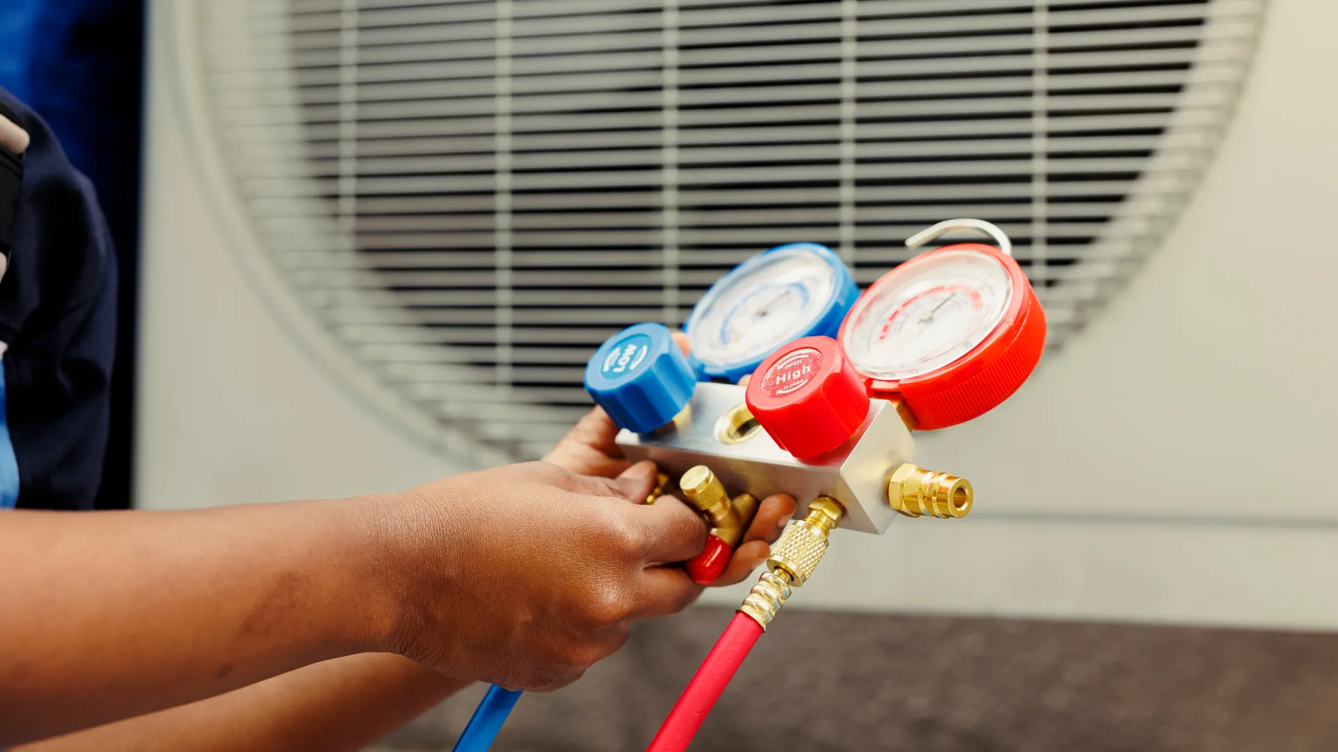Technician holding HVAC gauges with red and blue dials connected to air conditioning unit outdoor condenser.