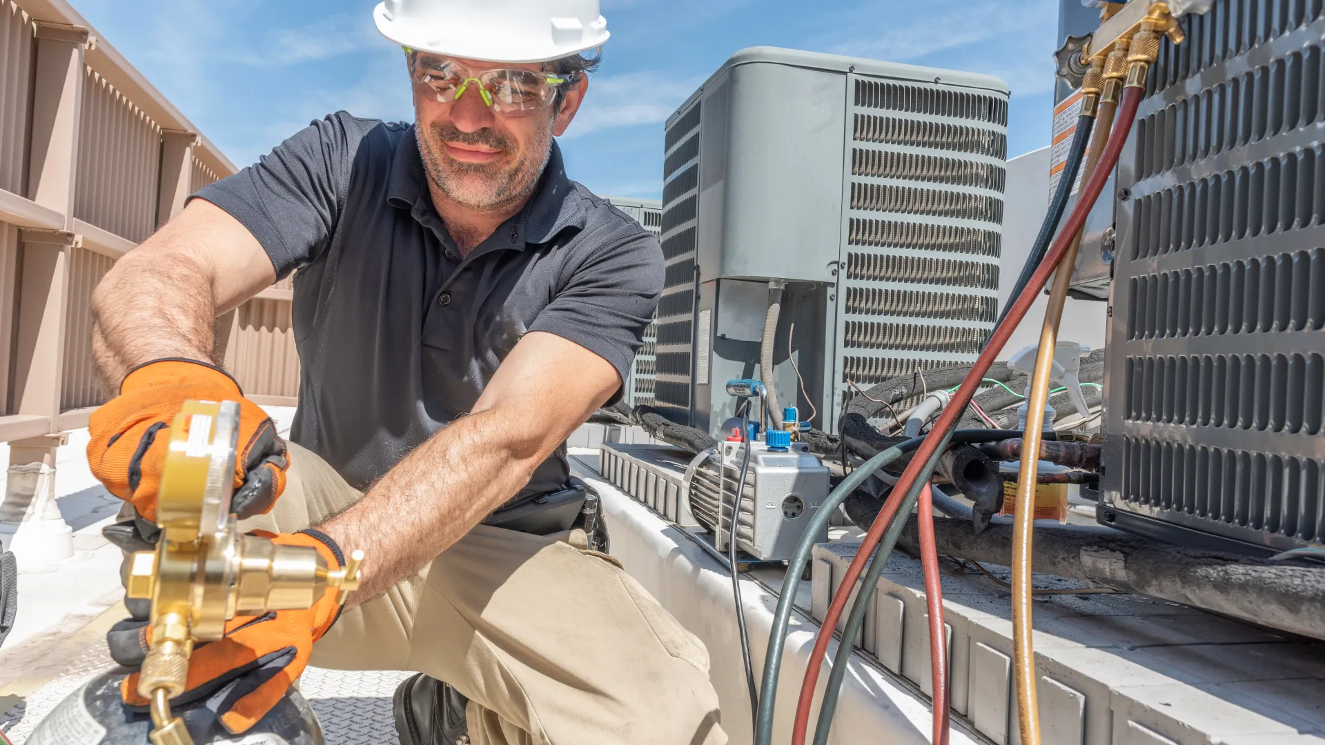 Technician wearing safety gear repairs rooftop air conditioning unit using gauges and refrigerant tanks under clear sky.