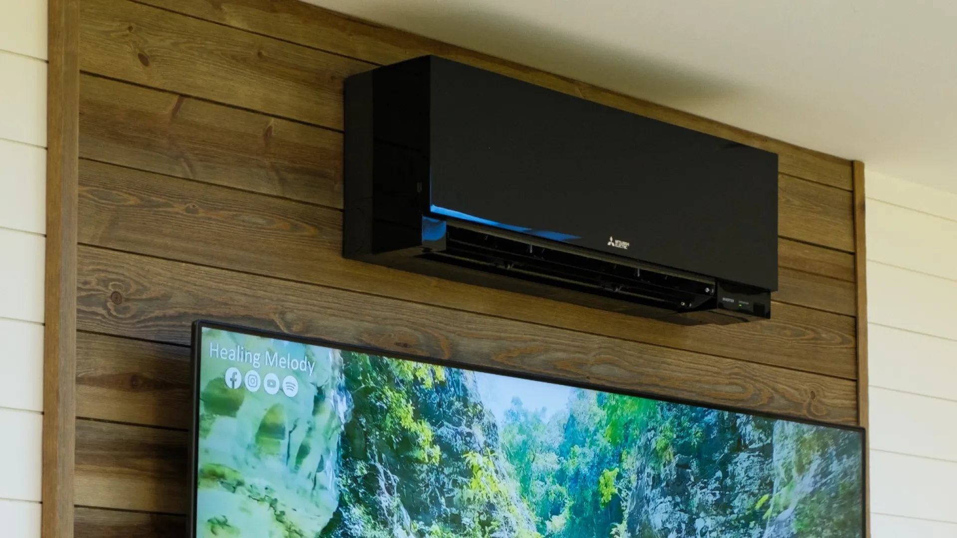 Flat-screen TV mounted on a wooden wall showing a peaceful river canyon scene beneath a black air conditioning unit.