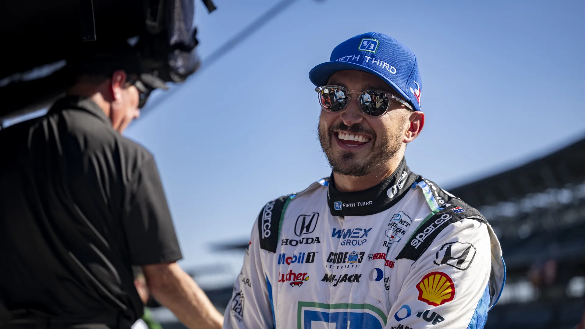 Smiling race car driver in sponsored fireproof suit and blue Fifth Third cap at racetrack under clear sky.
