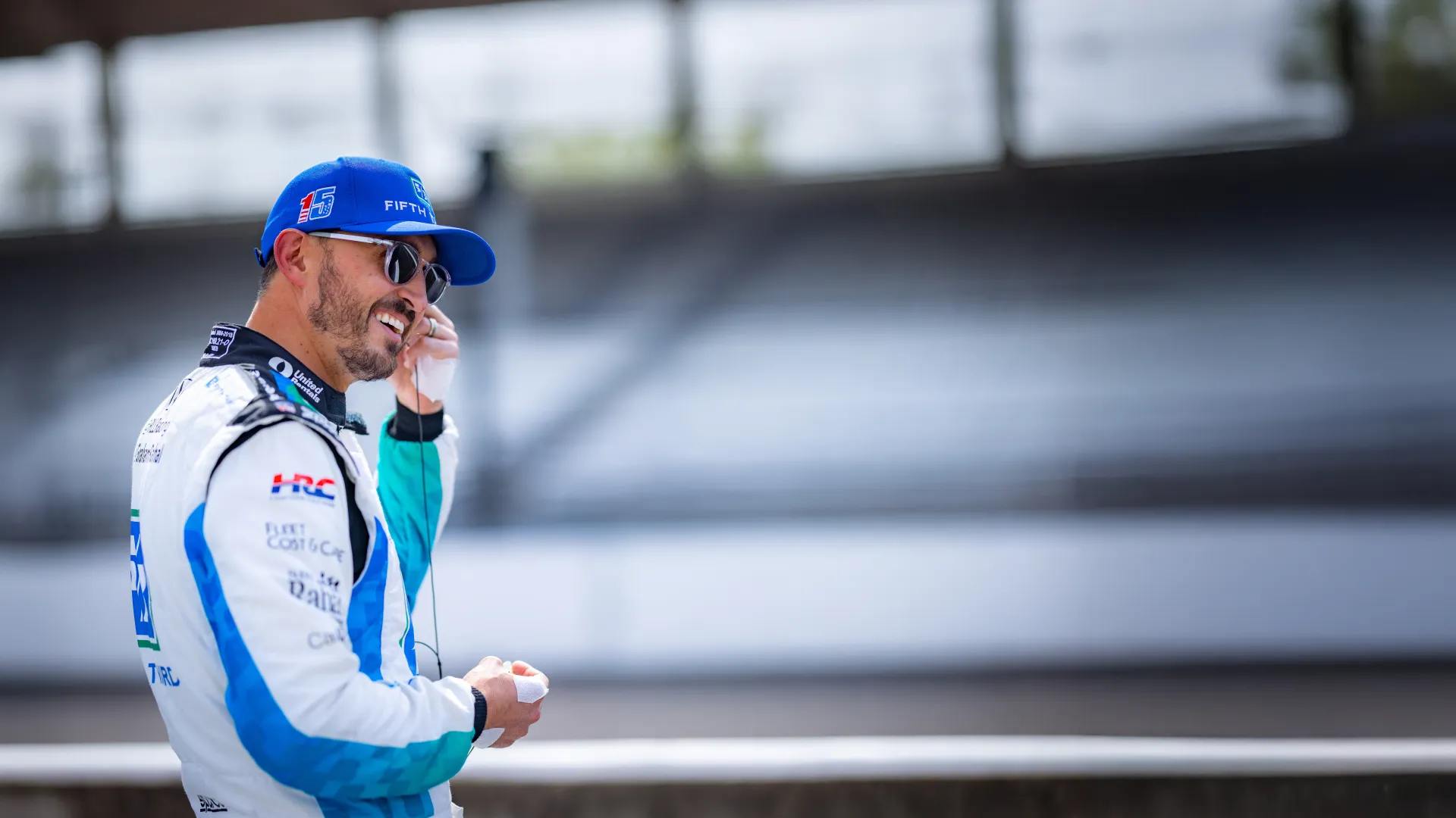 Racecar driver in white and blue suit adjusting earpiece in empty stadium with blurred background