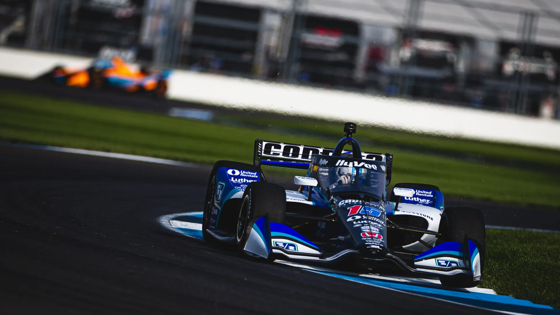 Blue and white IndyCar racing at high speed on a curved racetrack with another car in the background.