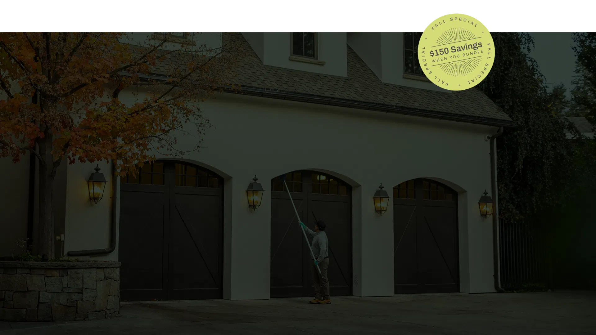 Man cleaning large white house garage doors with a long brush under warm wall lanterns in early evening.