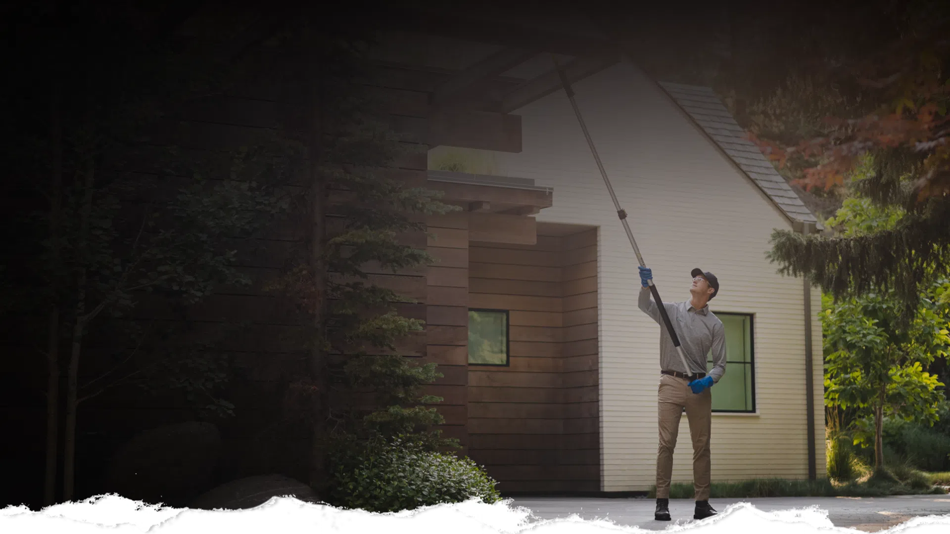 Man using a long pole to clean or inspect the exterior of a modern house surrounded by greenery.