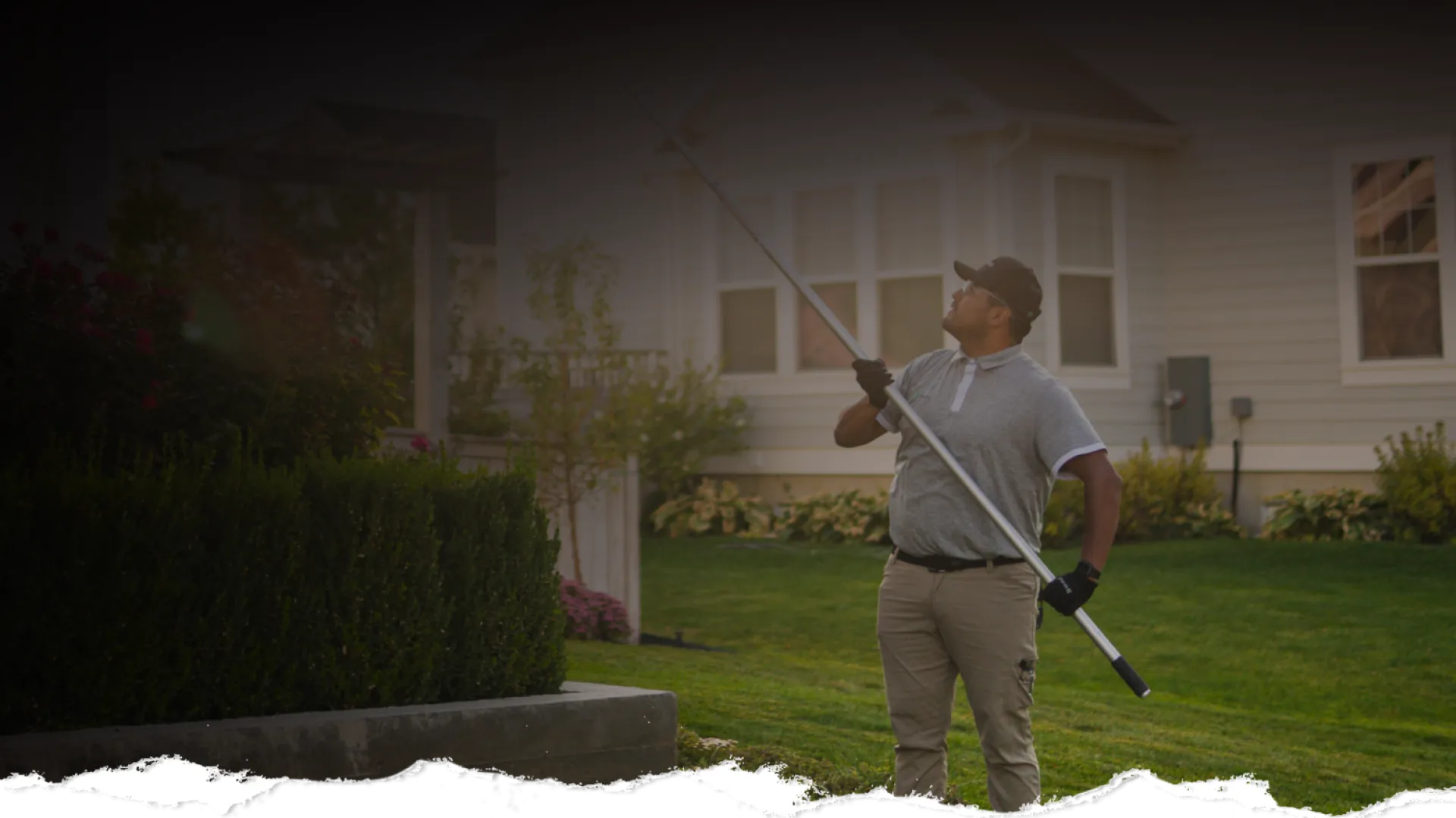 Man using a long pole to trim tall hedges in a residential garden on a sunny day.