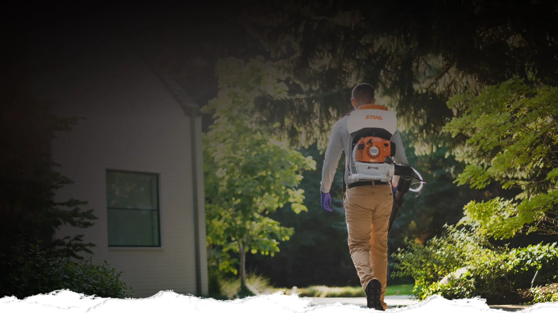 Person wearing a backpack leaf blower walking along a garden path near a house surrounded by green trees.