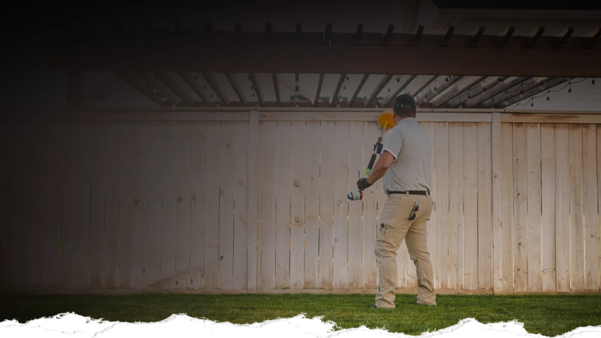Man in work clothes cleaning outdoor area with a long brush near a wooden fence under a pergola.