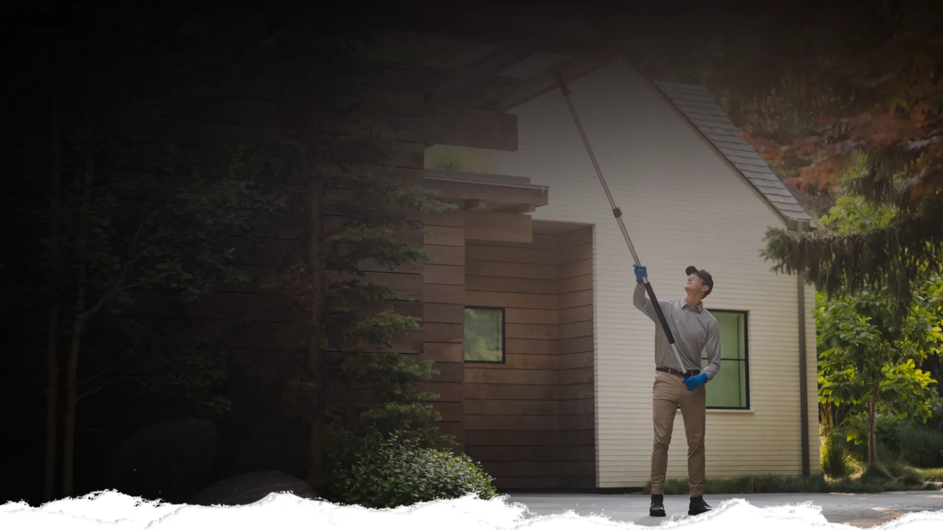 Person cleaning gutters on a house exterior using a long pole in a sunny outdoor setting
