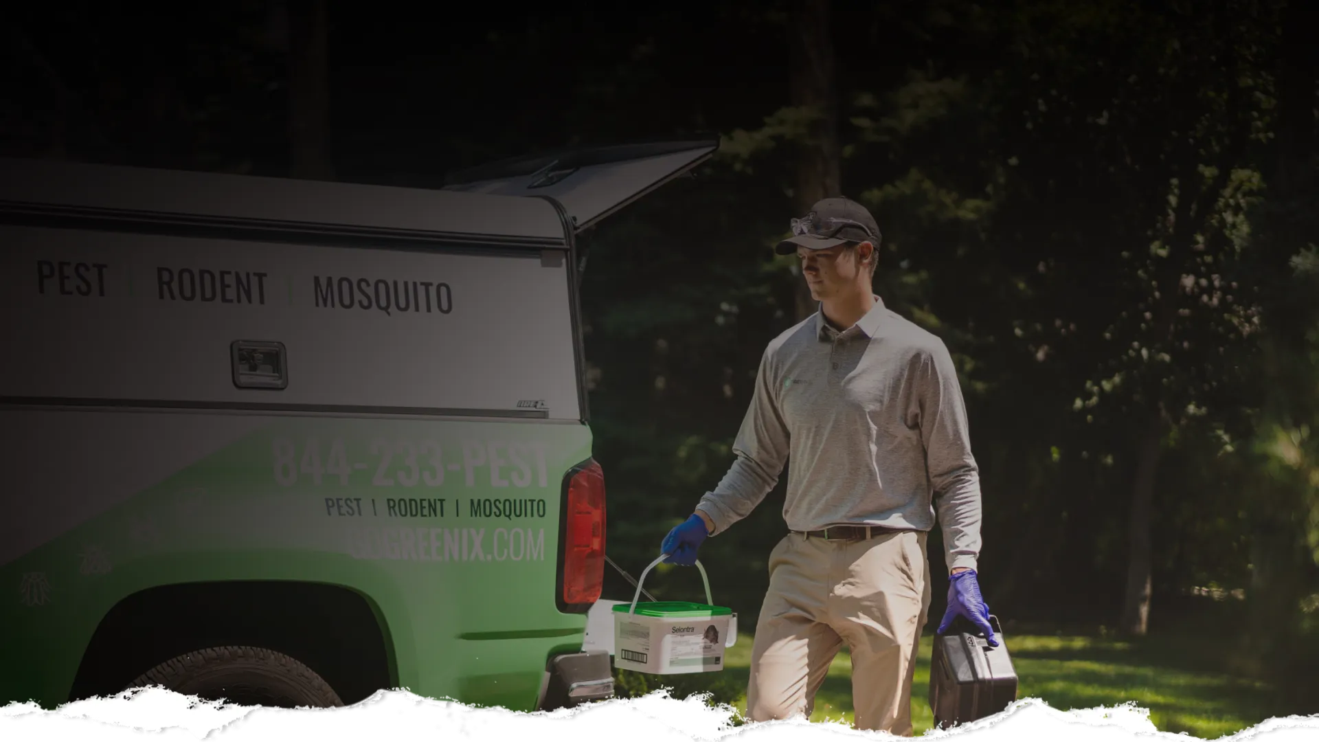 Pest control technician carrying equipment near a green and white truck in a wooded outdoor area.